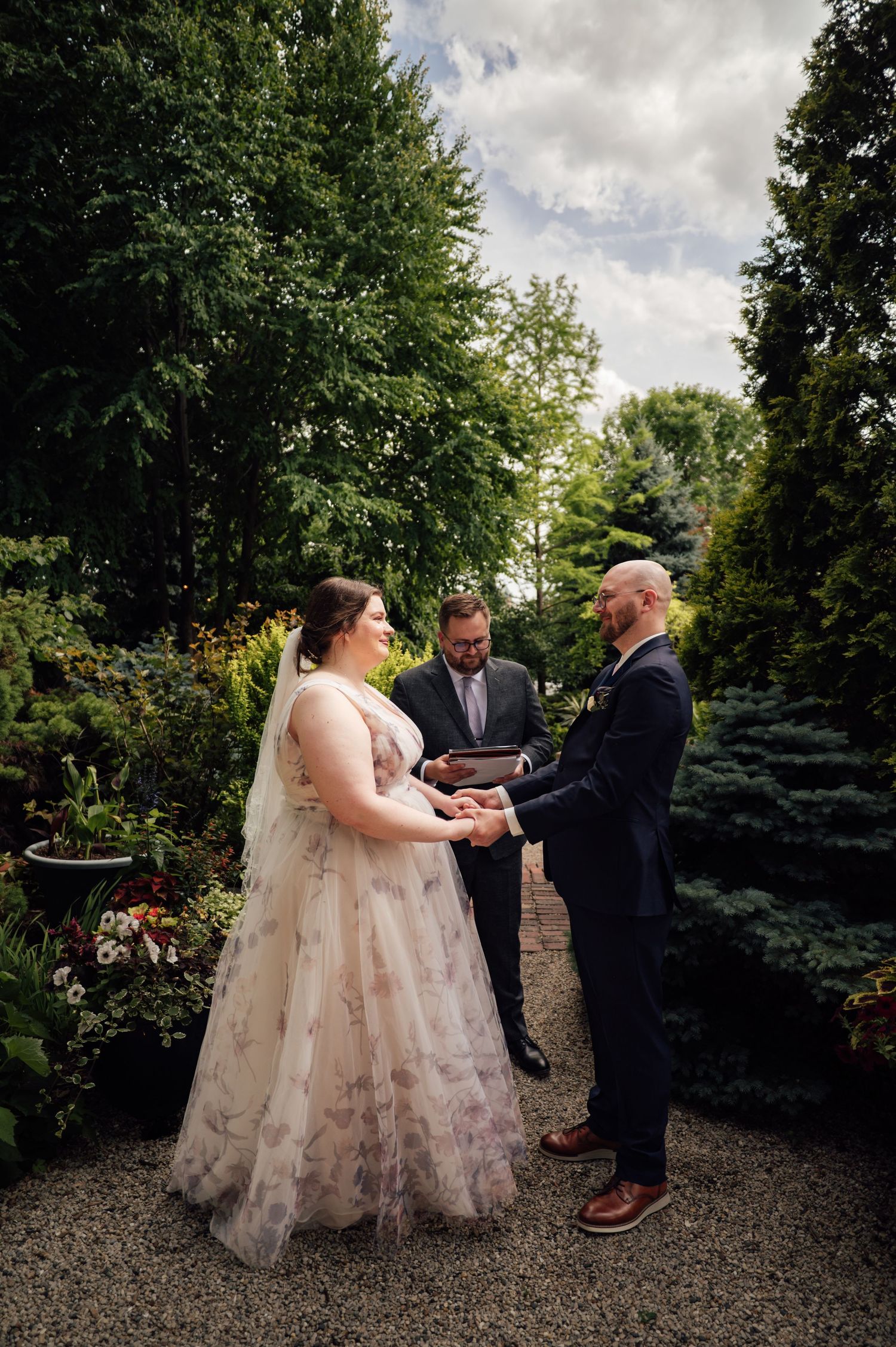 A couple exchanges vows in a lush garden ceremony, bride in floral gown with veil, groom in navy suit, officiant behind them.