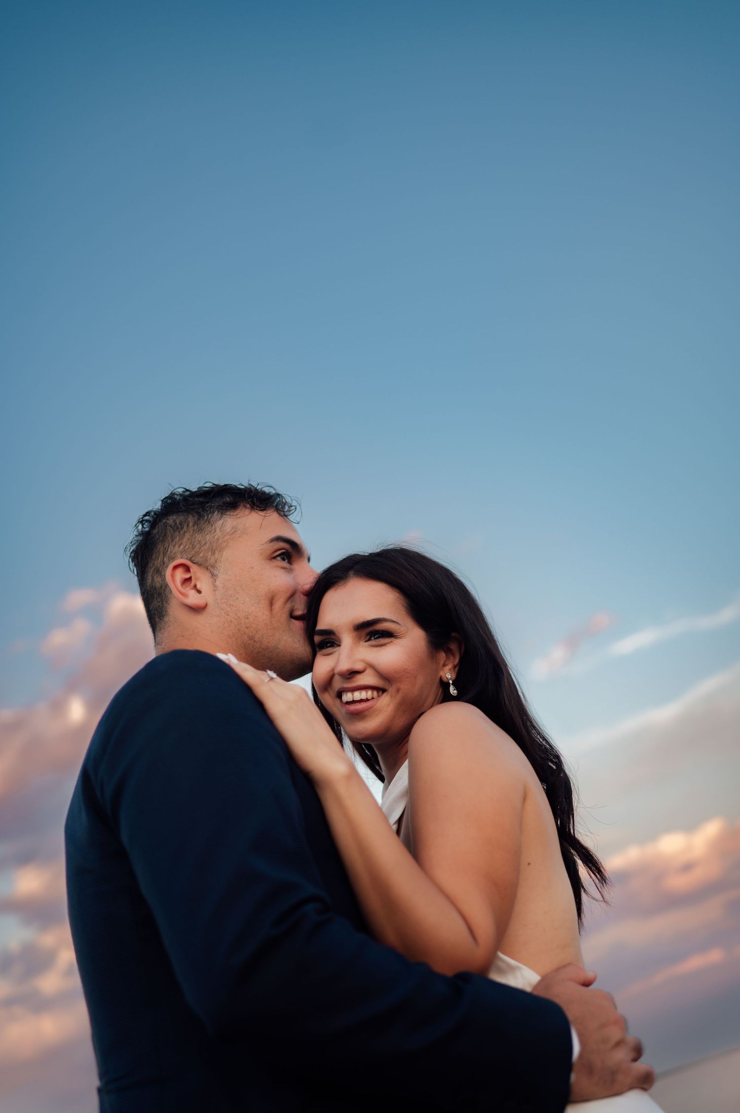 A couple embraces at sunset, the bride smiling joyfully as the groom whispers in her ear against a blue sky.