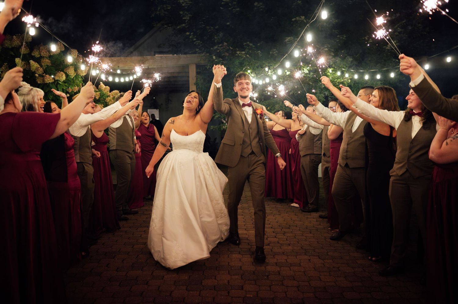 Bride and groom walk through sparkler exit surrounded by guests in burgundy dresses at outdoor night wedding celebration.
