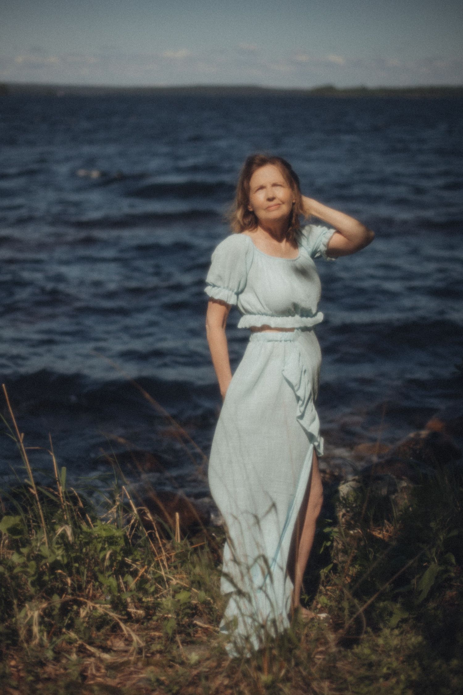 Nainen seisomassa meren rannalla, tuuli ja luonnonvalo luovat rauhallisen hetken