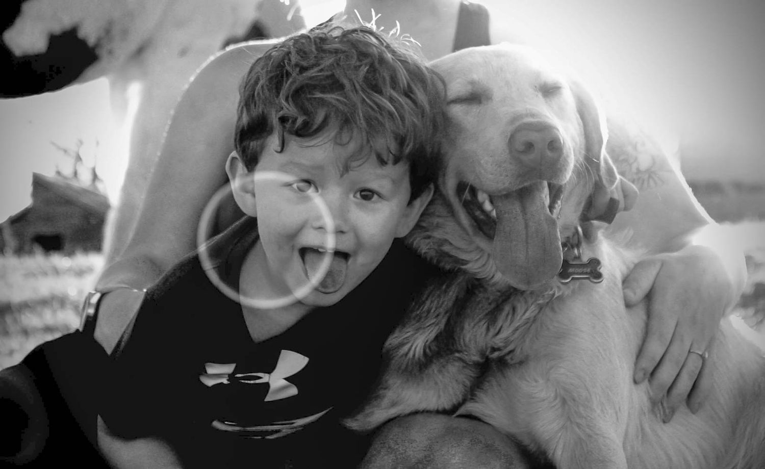 Black and white photo of a laughing boy hugging a happy Labrador dog outdoors.