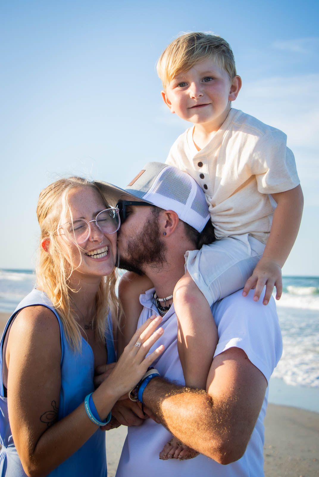 Family of three enjoying a beach day, parents smiling and kissing while young blonde boy sits on dad’s shoulders.