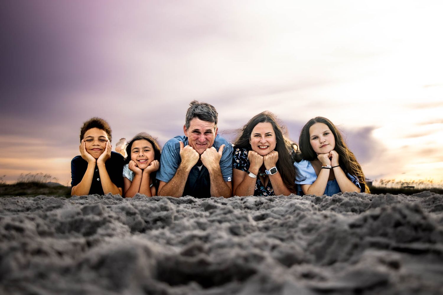 Family of five lying on sandy beach at sunset, smiling at camera with colorful sky backdrop.