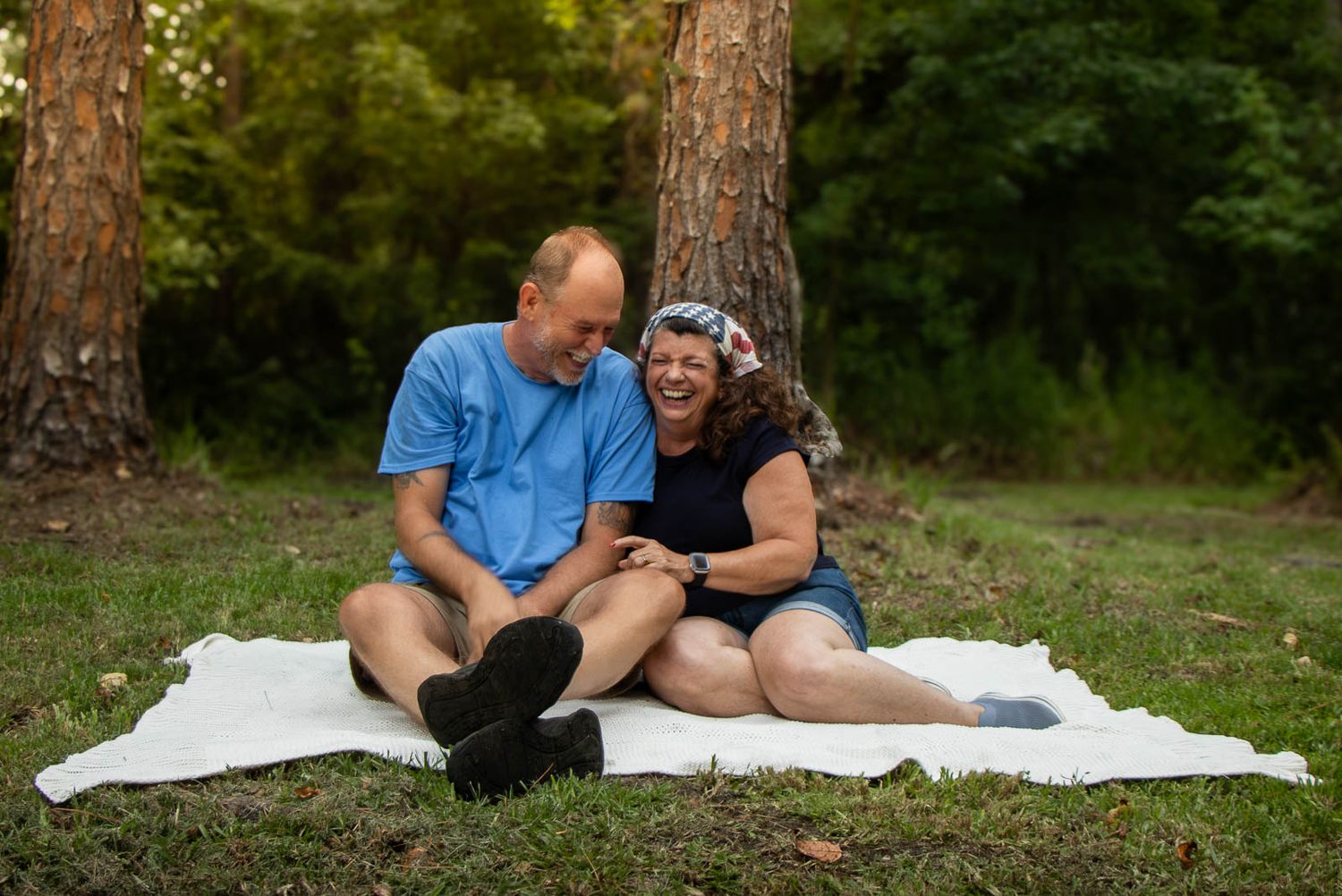 Couple laughing together on white blanket in park, sitting against trees on green grass on sunny day.