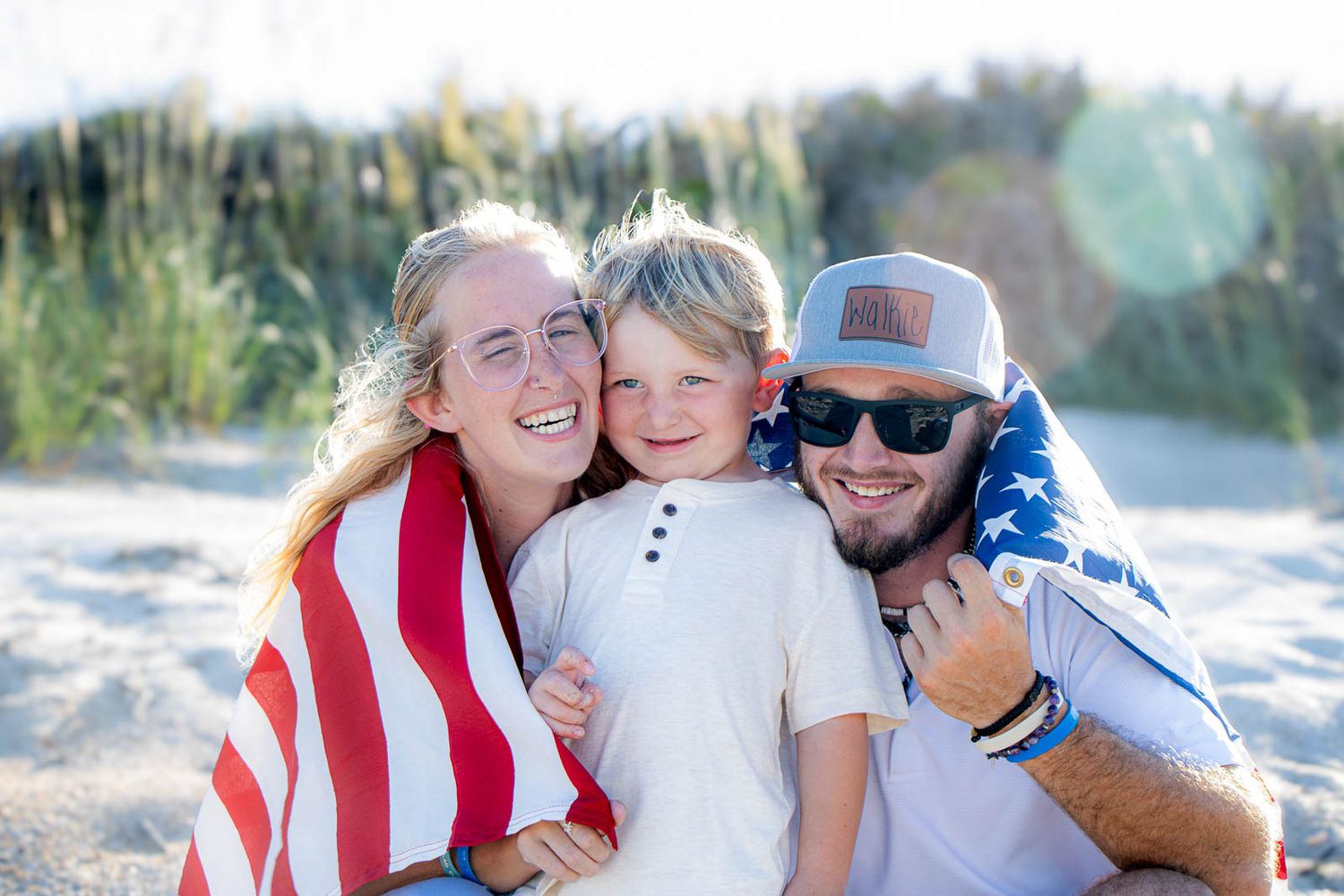 Happy family of three wrapped in American flag smiling on sandy beach on a sunny day.