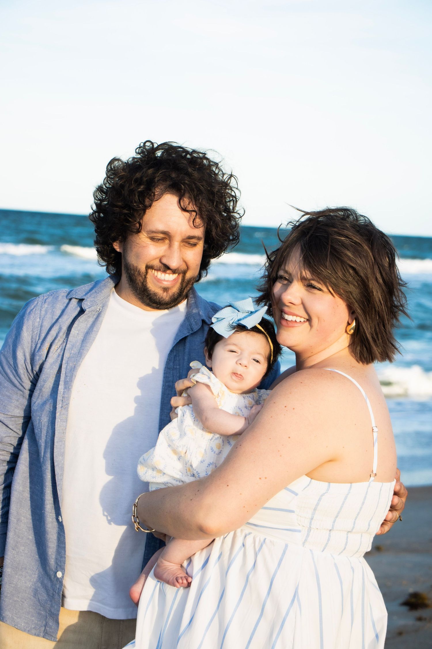 Happy couple smiling at their baby girl with blue bow on sunny beach shoreline.
