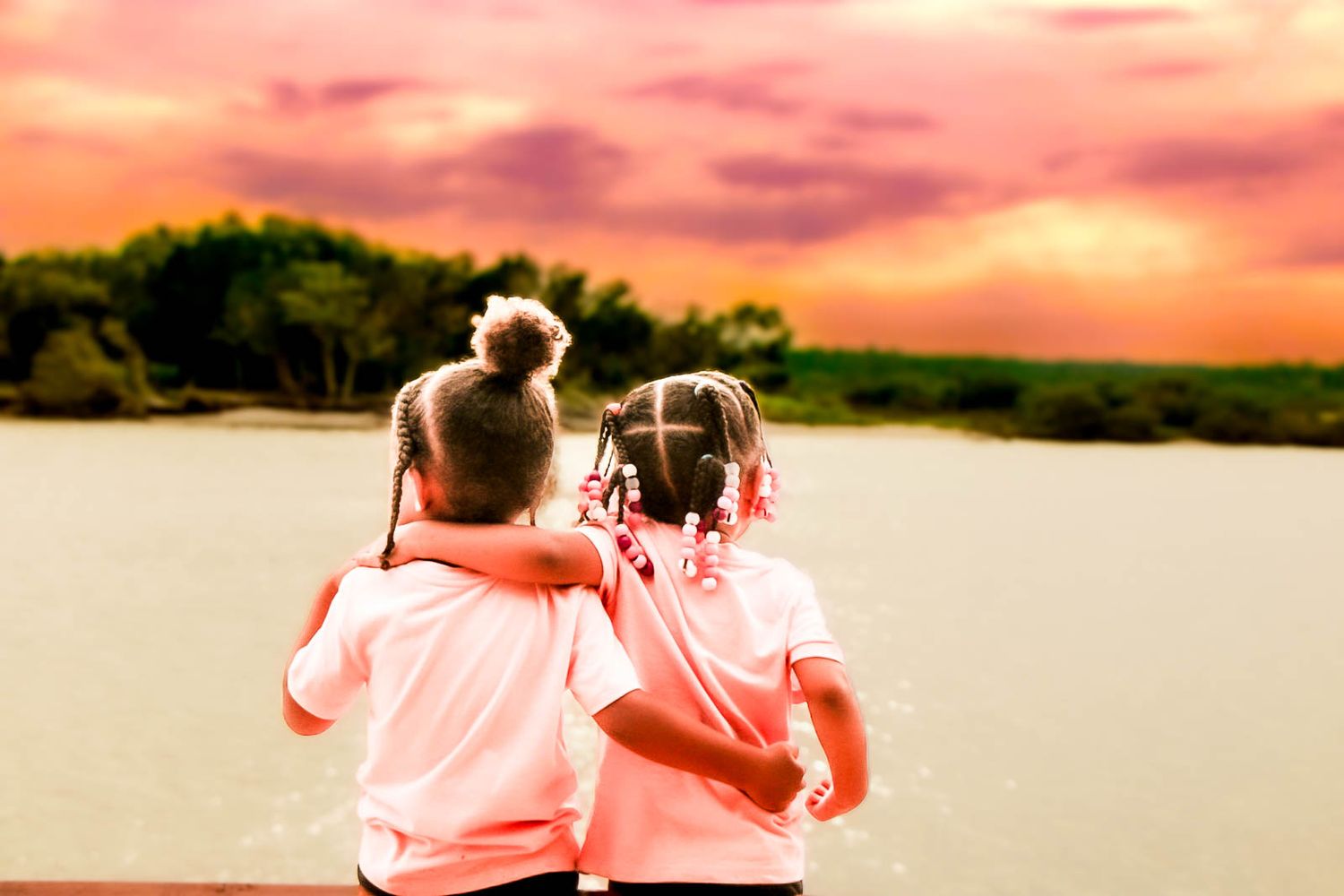 Two young girls with buns embrace while gazing at a scenic lake under a vibrant pink and orange sunset sky.