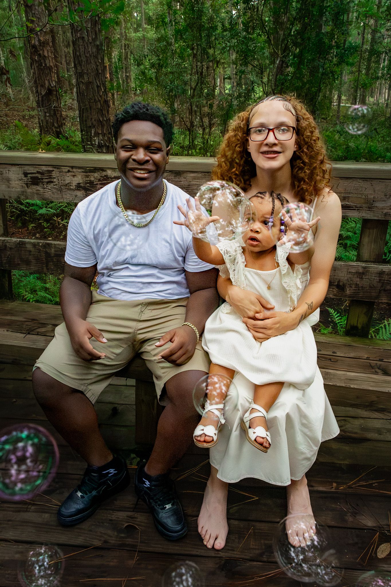 Interracial family with baby girl sitting on wooden deck surrounded by trees, playing with bubbles during outdoor photoshoot.