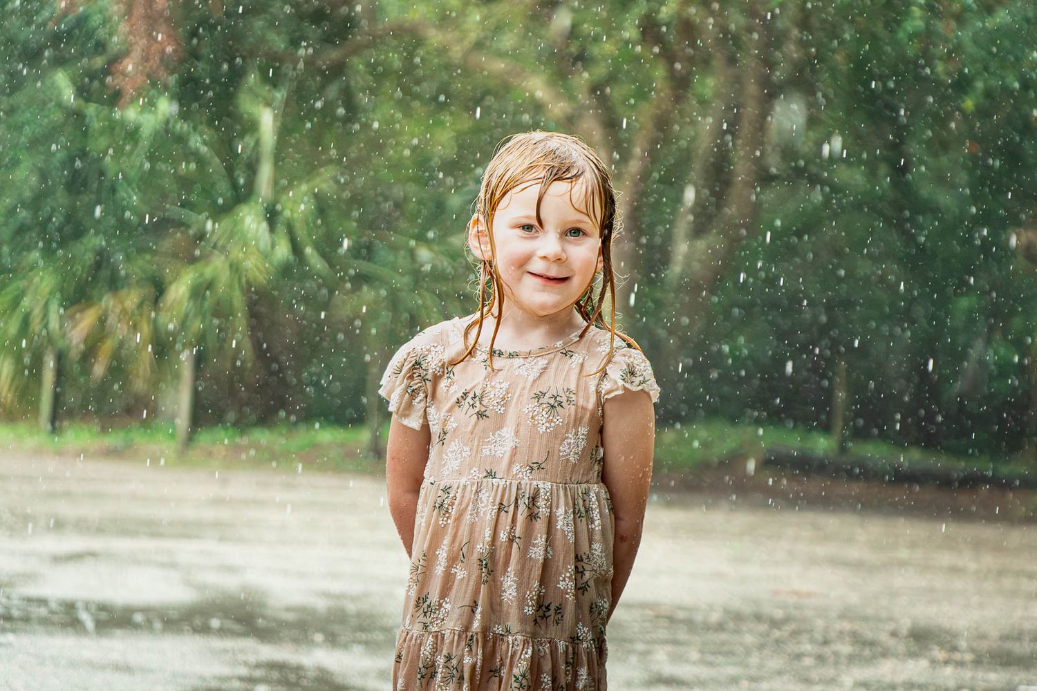 A smiling little girl in a floral dress stands happily in the rain surrounded by lush green trees.