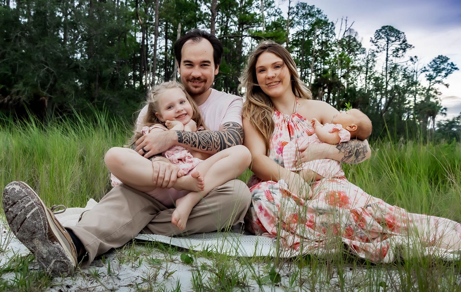 Family of four sitting in green meadow with parents holding young daughter and newborn baby for family photos in Jacksonville, Florida.
