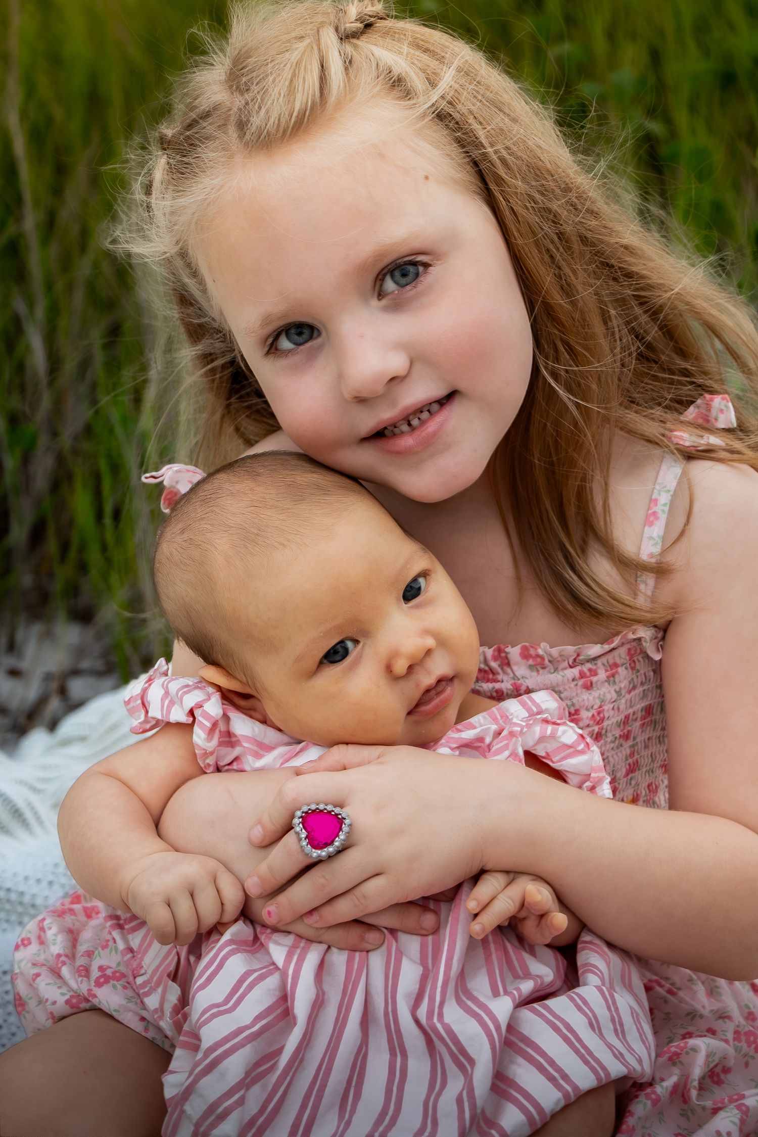 Young blonde girl in pink dress holds infant sibling outdoors near green grass, wearing a pink heart ring.