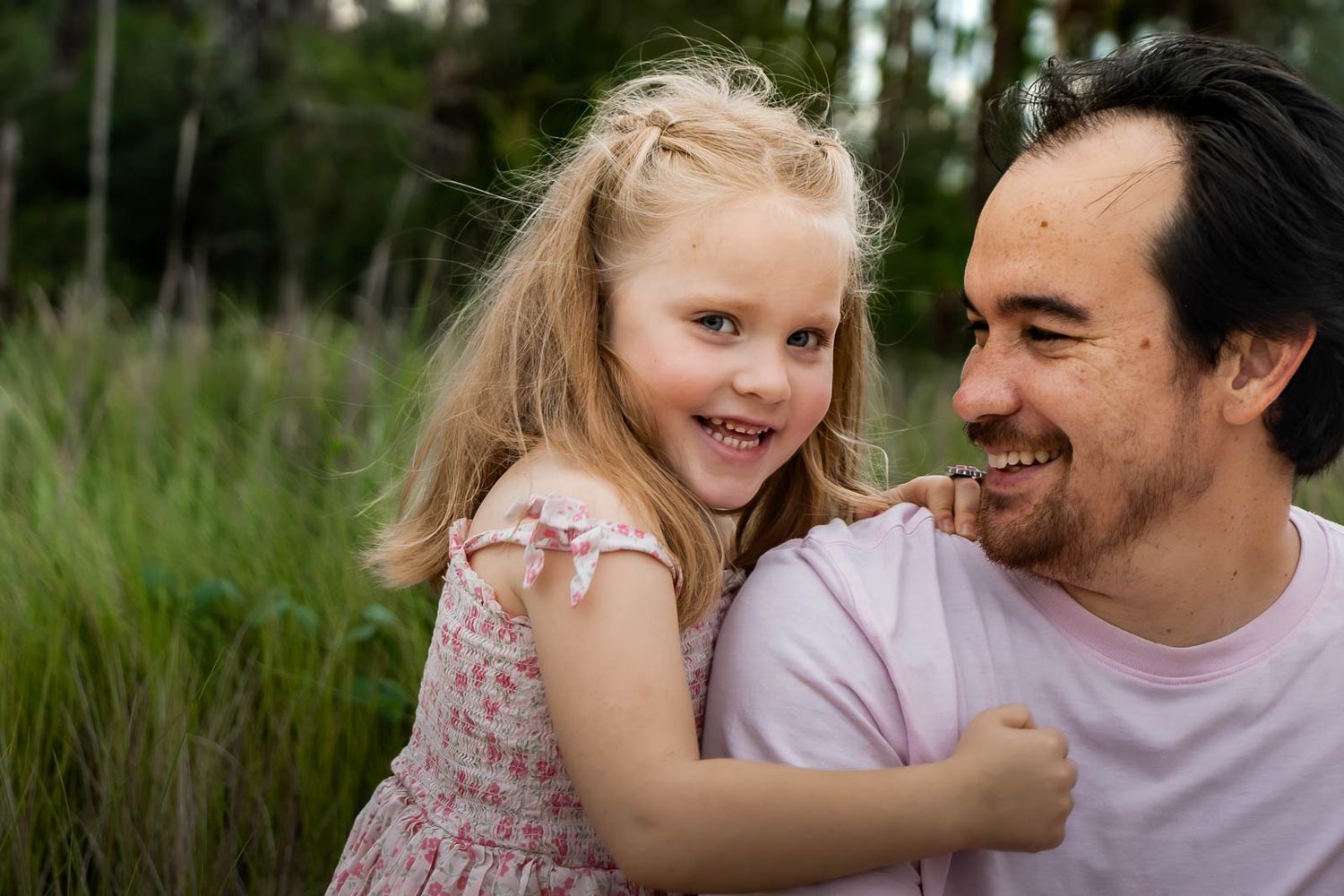 A smiling young blonde girl hugs a laughing dark-haired man outdoors in a lush green field.