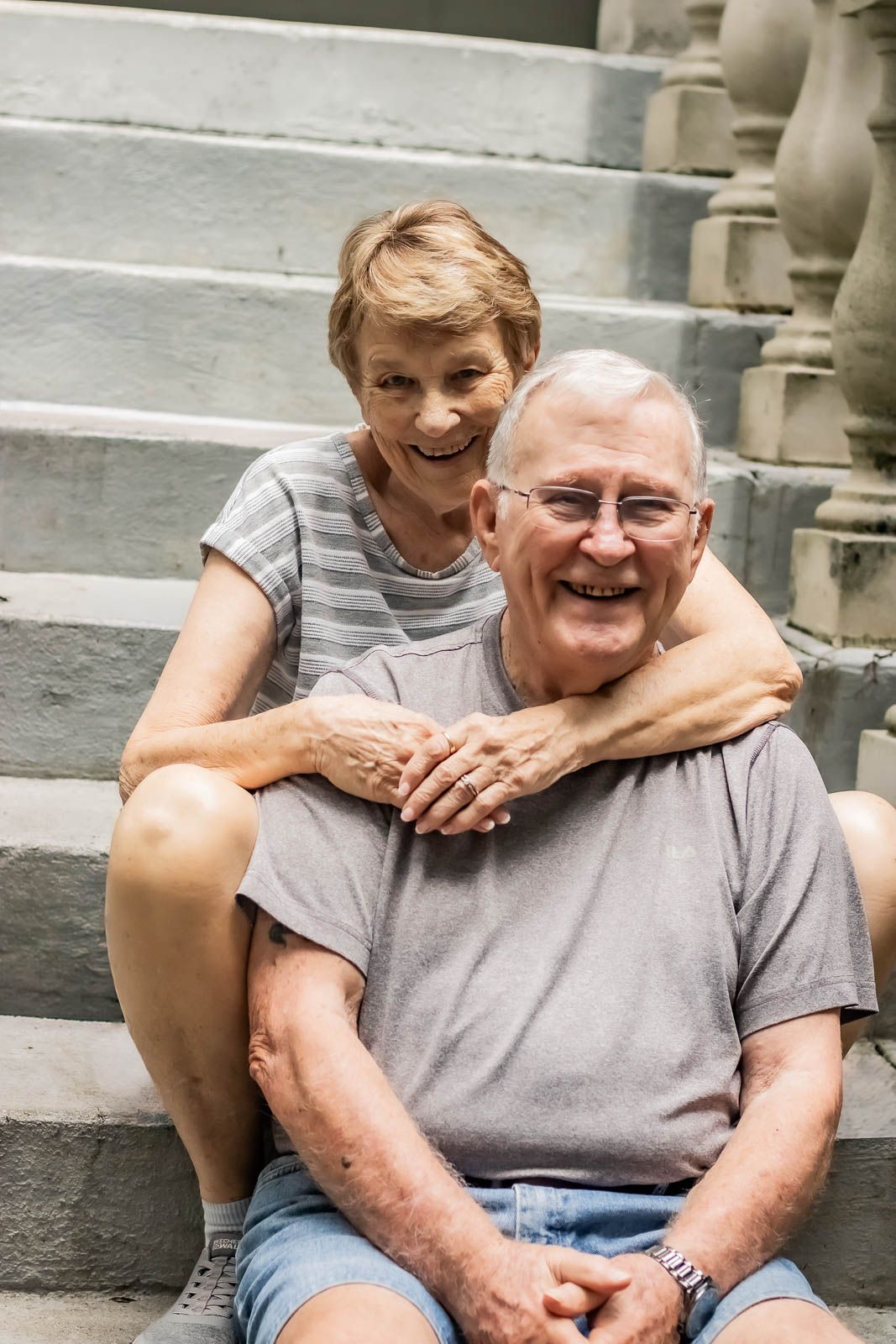 Happy senior couple sitting on stone steps, smiling warmly, woman in casual summer attire during their portrait session in Orange Park, fl.