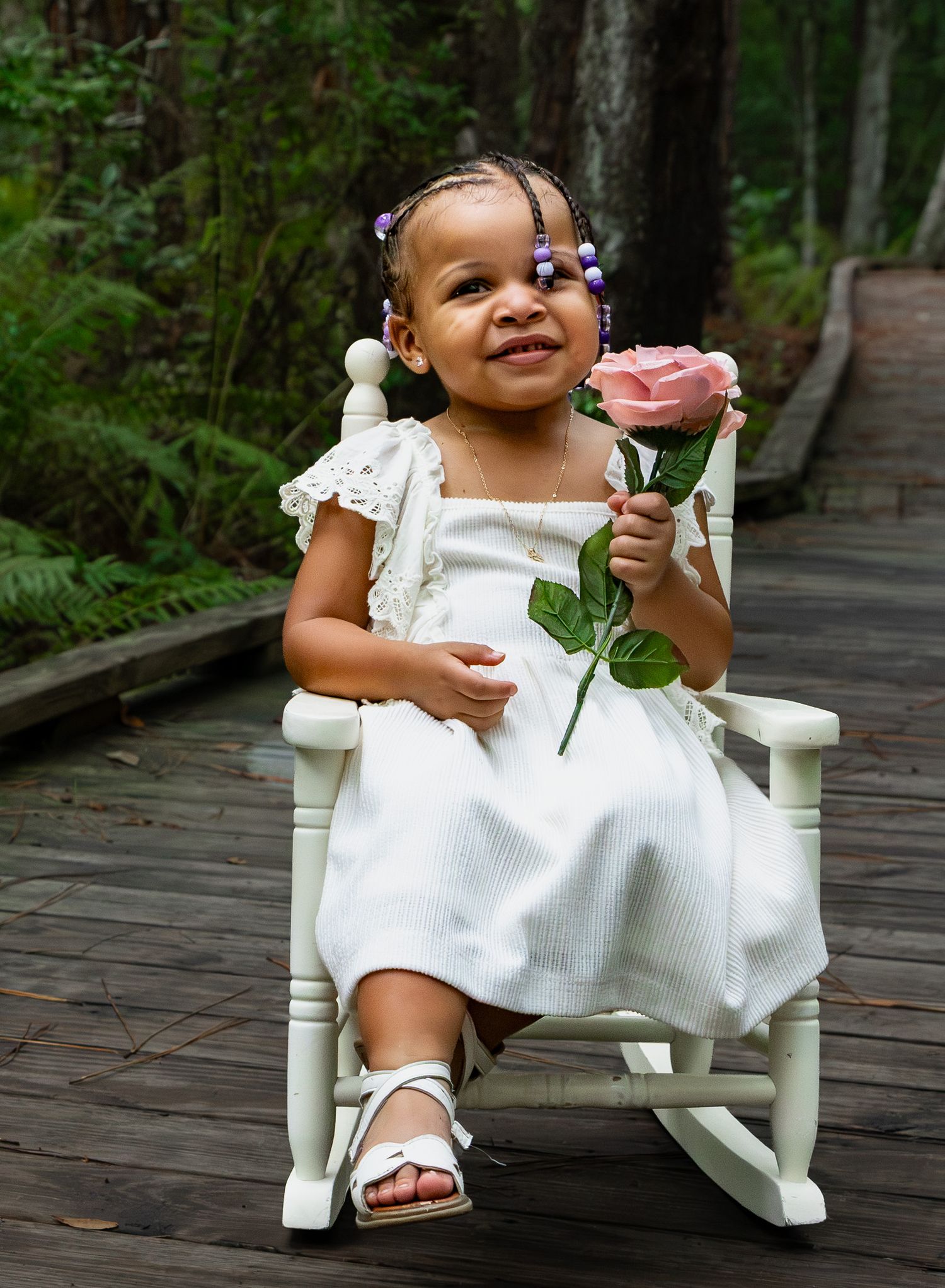 Smiling toddler girl in white dress holding a pink rose while sitting in a small rocking chair on a wooden boardwalk.