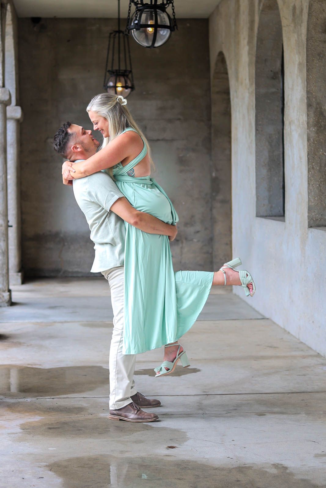 A man lifts a woman in a mint green dress as they share a joyful embrace in an elegant corridor.