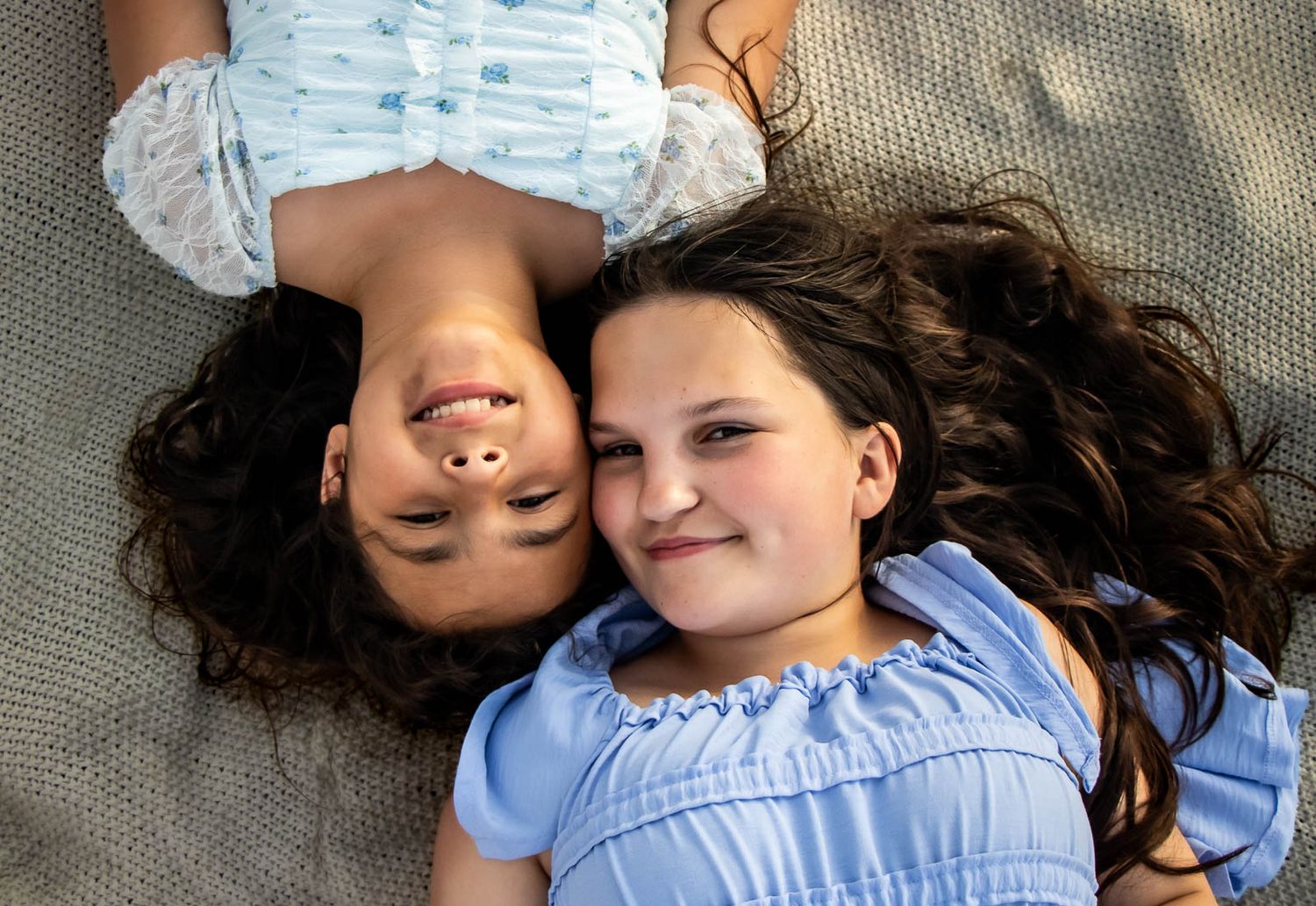 Two young girls with long dark hair lying down together smiling, wearing blue dresses outdoors.