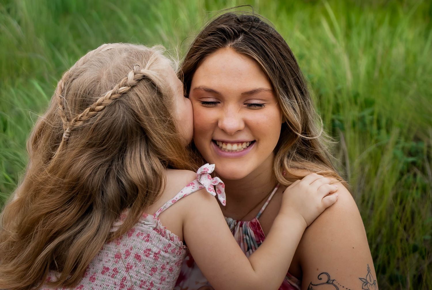A young girl kisses her smiling mother on the cheek in a lush green field during a family photo session in Jacksonville, florida.