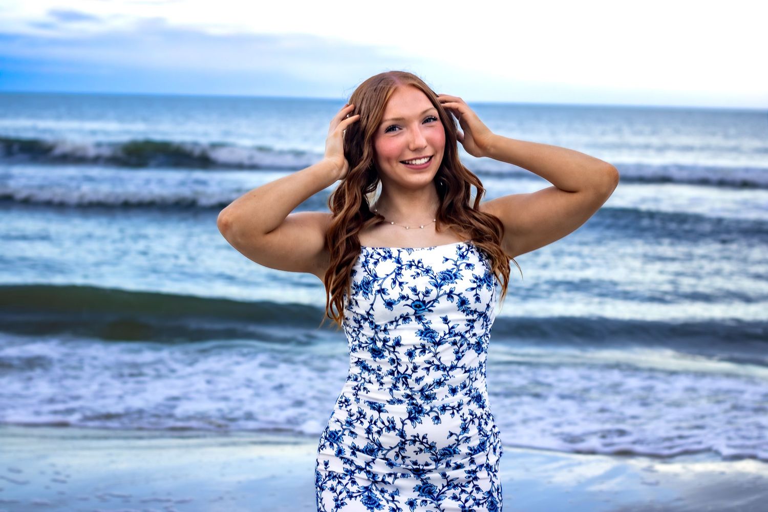 Young woman in white floral sundress smiling at beach, hands in hair, ocean waves in background.