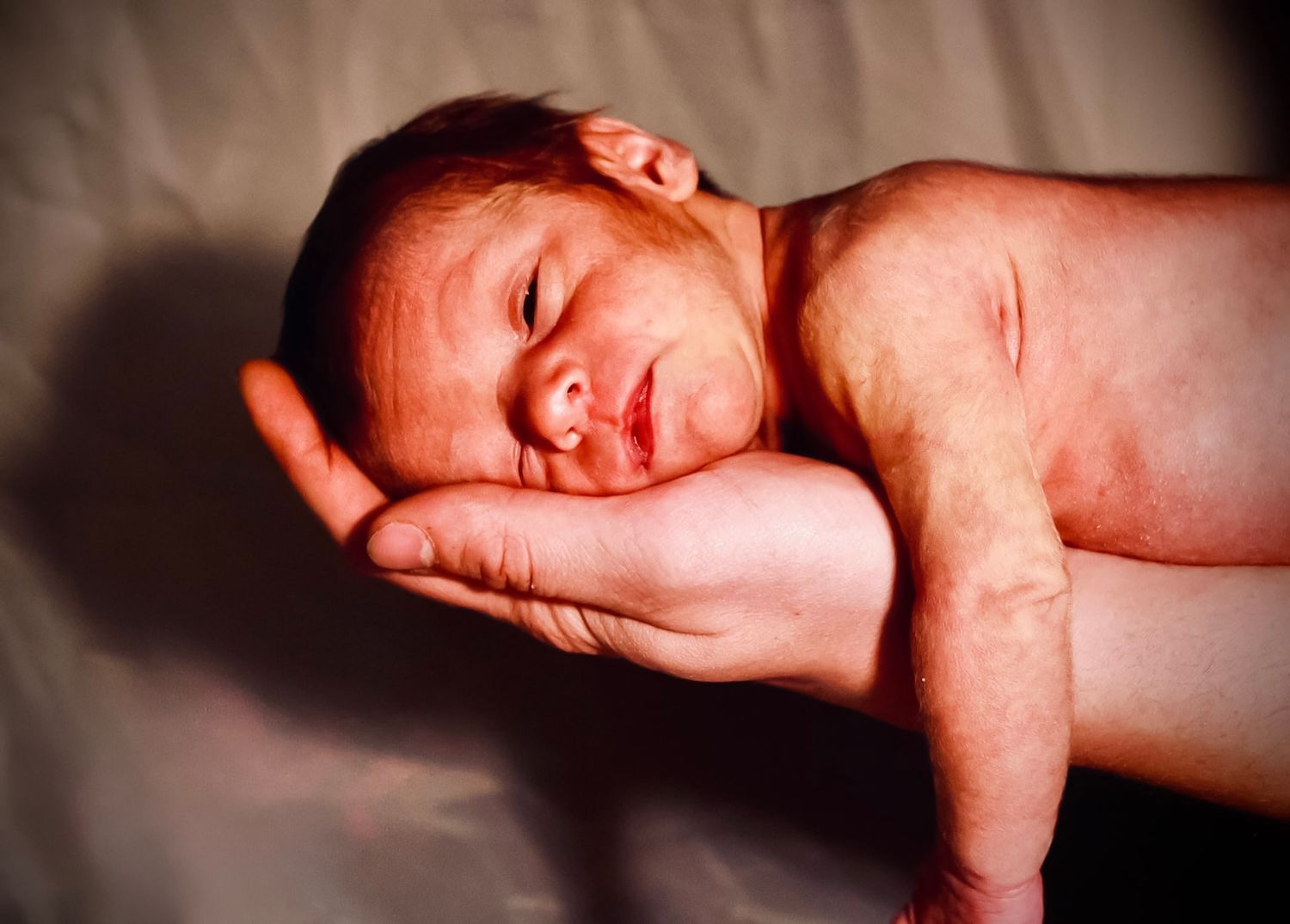 A newborn being help by his father looking at the camera with a gray background.
