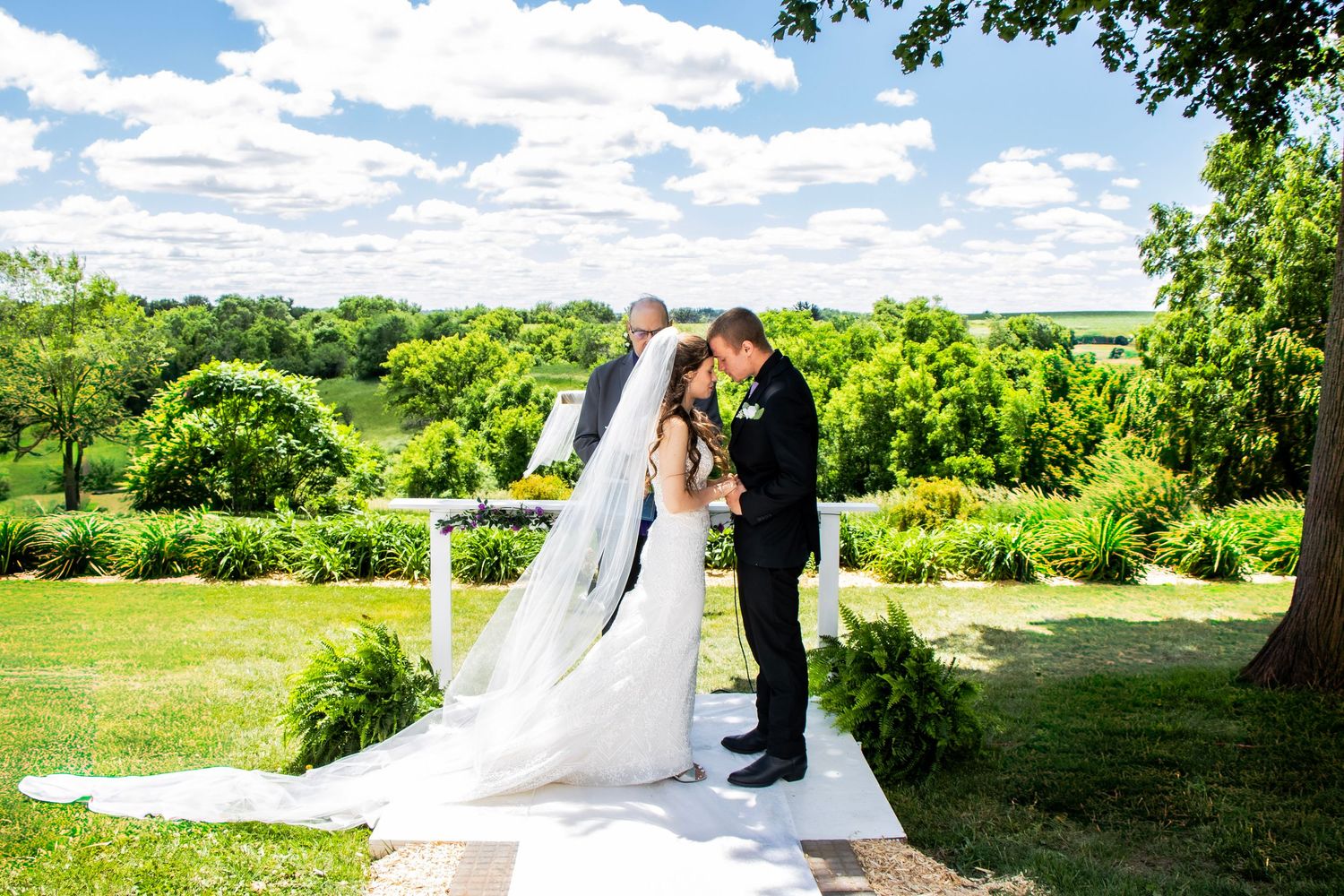 A man and a women during their wedding ceremony saying a prayer touching their foreheads together with vibrant greens in the background.