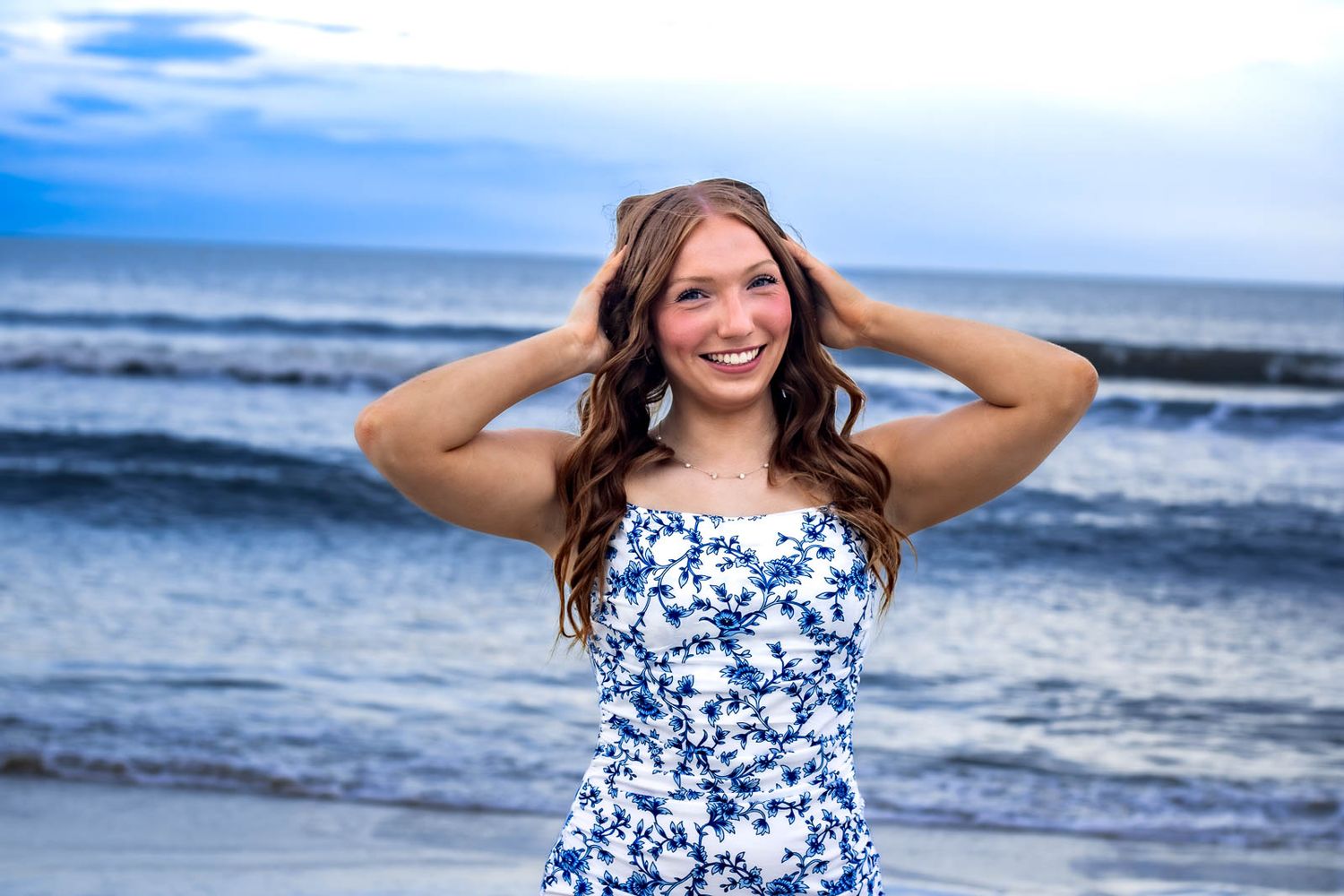 Young woman in blue floral dress smiling at the beach with ocean waves in the background.