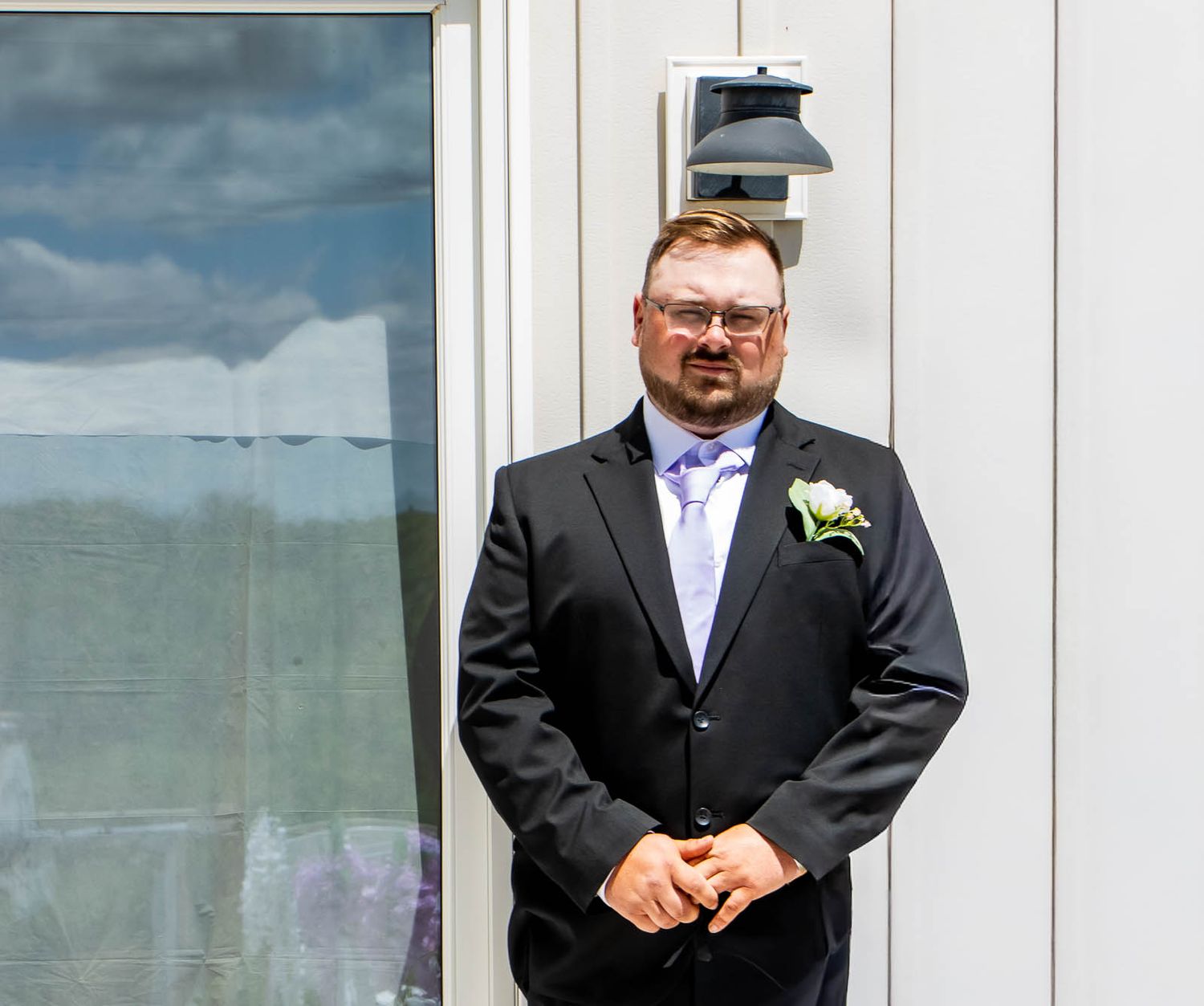 Man in black suit with boutonniere and lavender tie standing outdoors at a wedding ceremony.