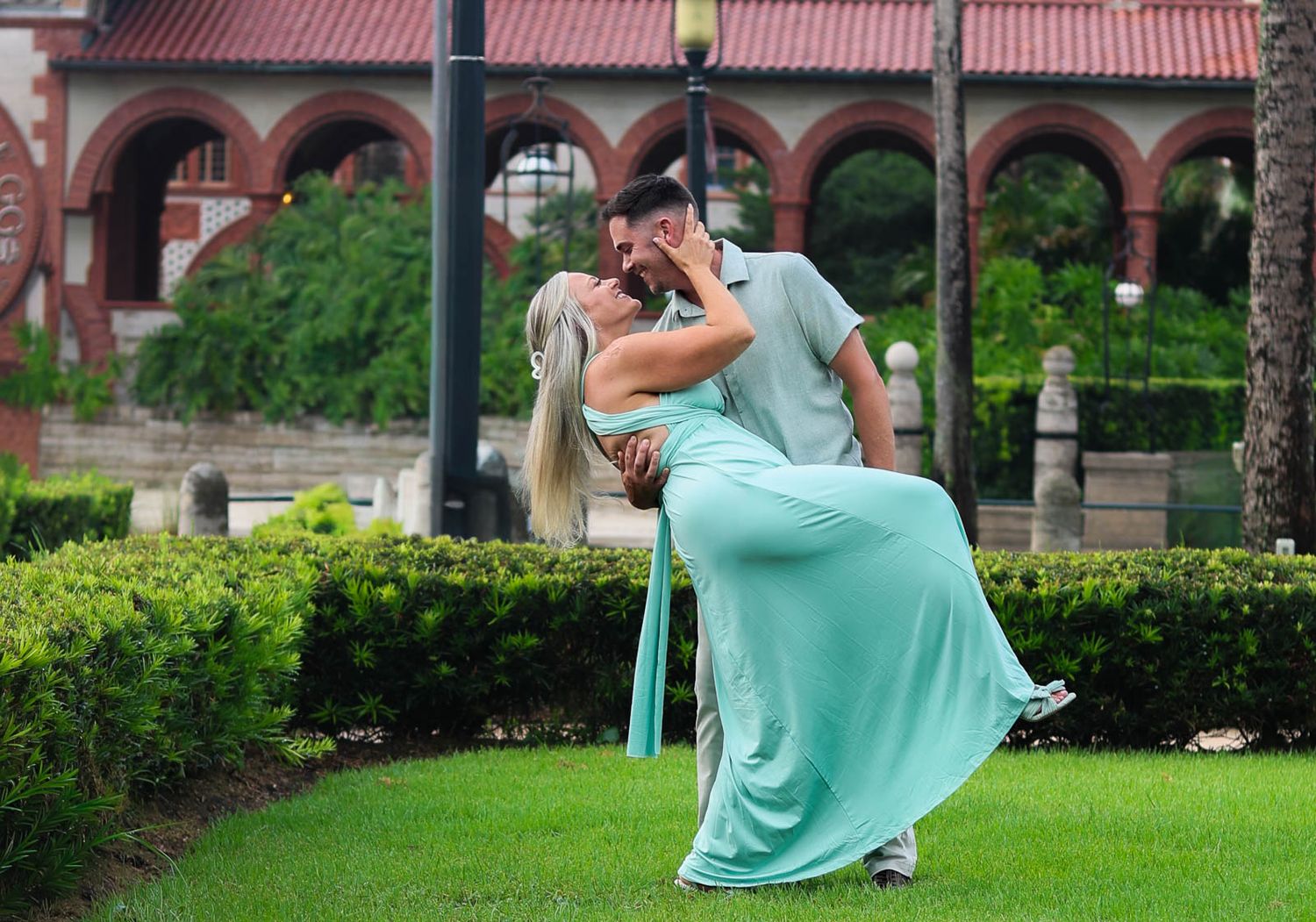 Couple sharing a romantic dip kiss outdoors, woman in mint green gown, lush garden and historic building backdrop.