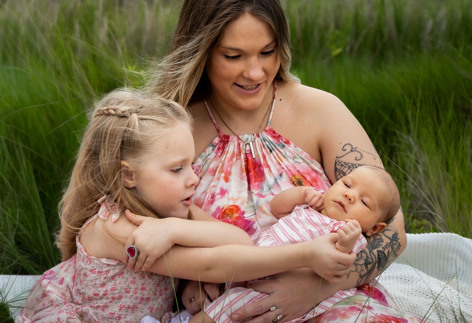 A tattooed mother in a floral dress holds her newborn while her young blonde daughter lovingly embraces them outdoors.