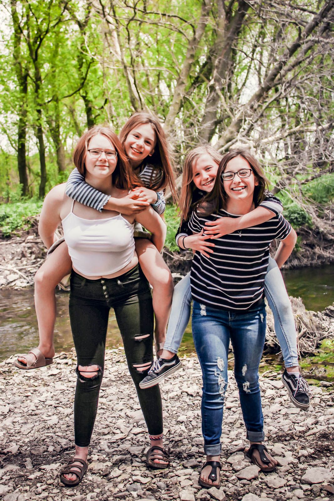 Four teenage girls enjoy piggyback rides outdoors near a rocky creek surrounded by lush green trees.