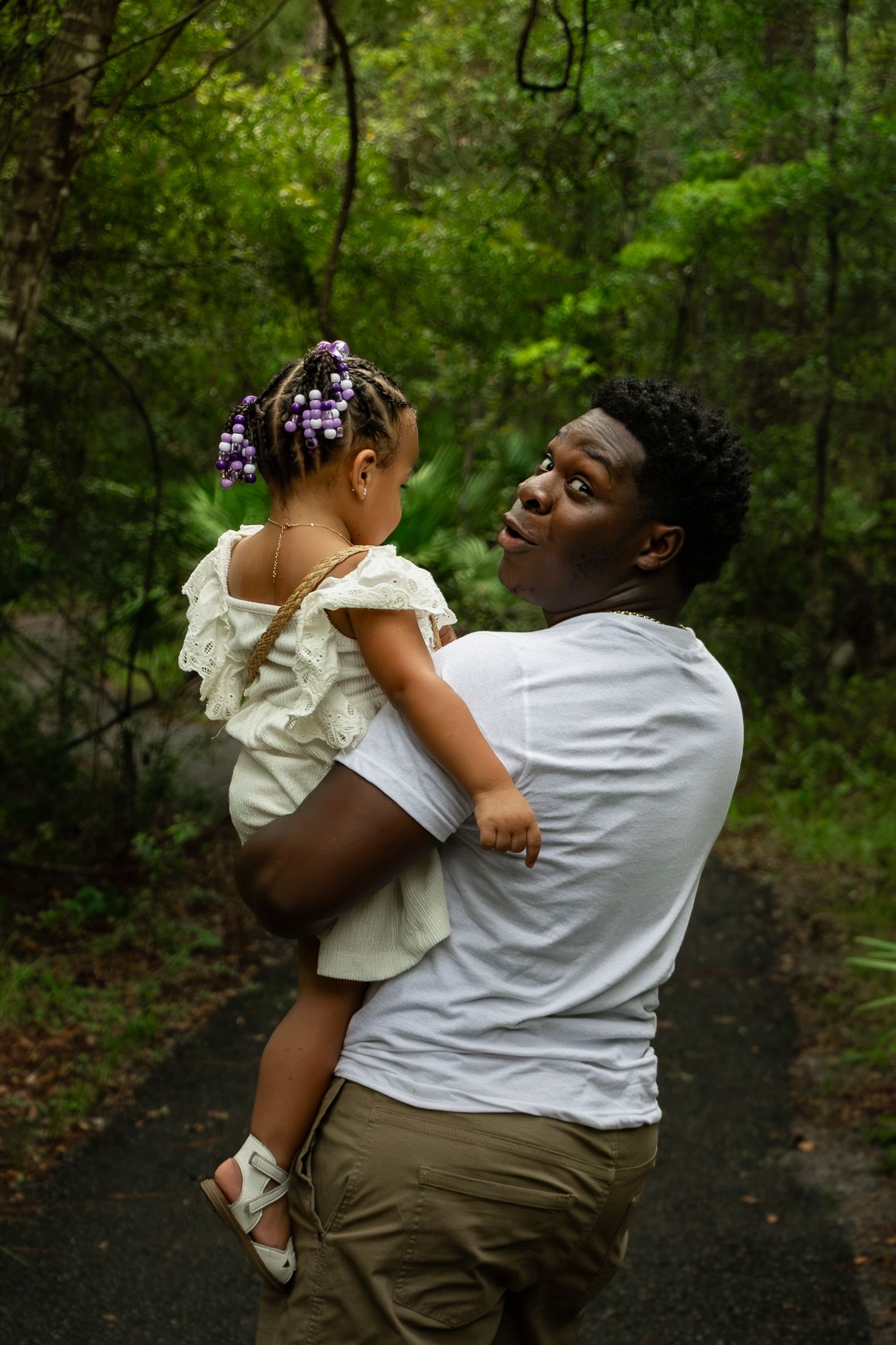 A man in a white shirt holds a young girl with purple hair accessories on a forest path.