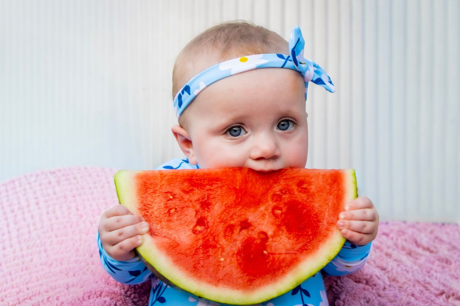 A baby sitting on a pink blanket eating a piece of red watermelon.