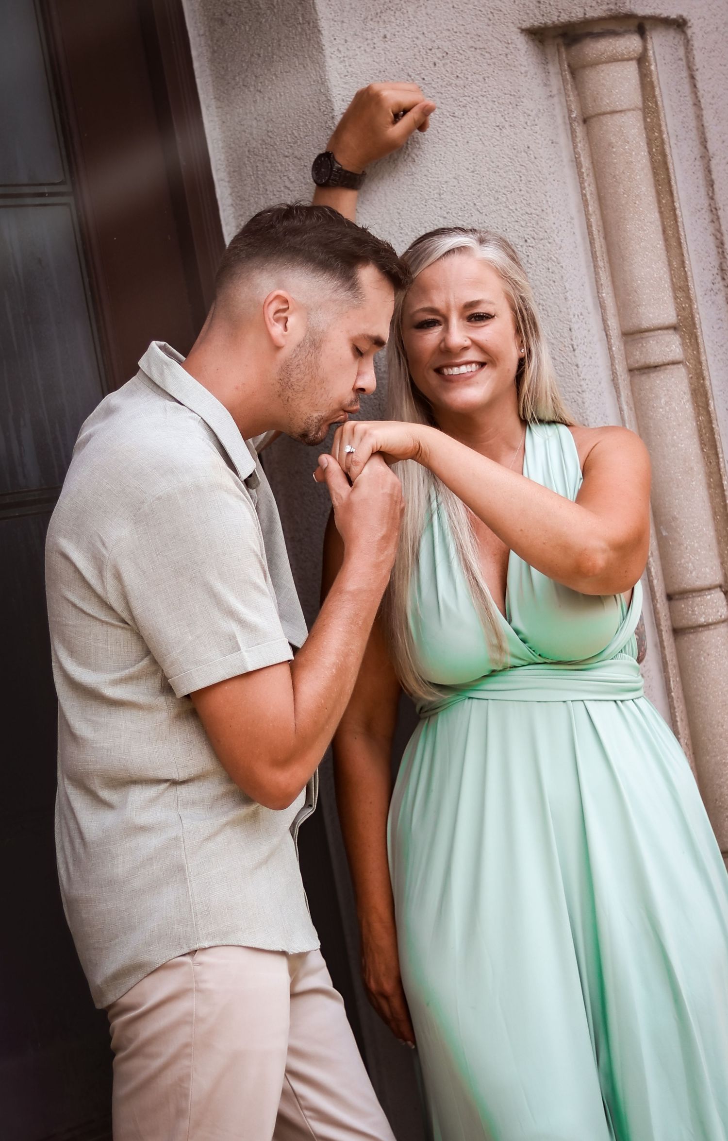 A man in beige kissing a smiling woman's hand in a mint green dress near a stone building entrance.