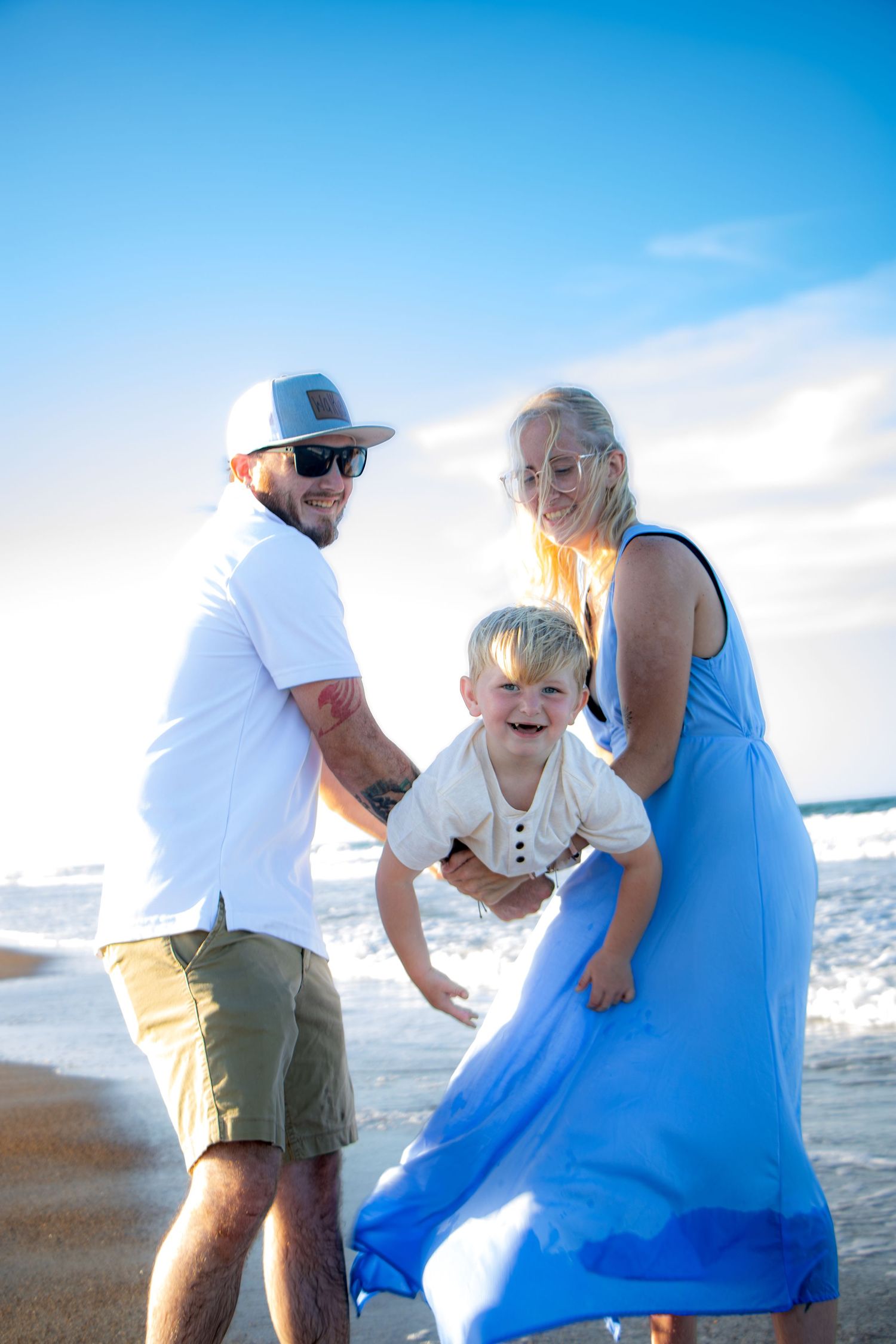 Happy family with young boy playing on sunny beach, waves in background, mother in blue dress and father in white shirt.