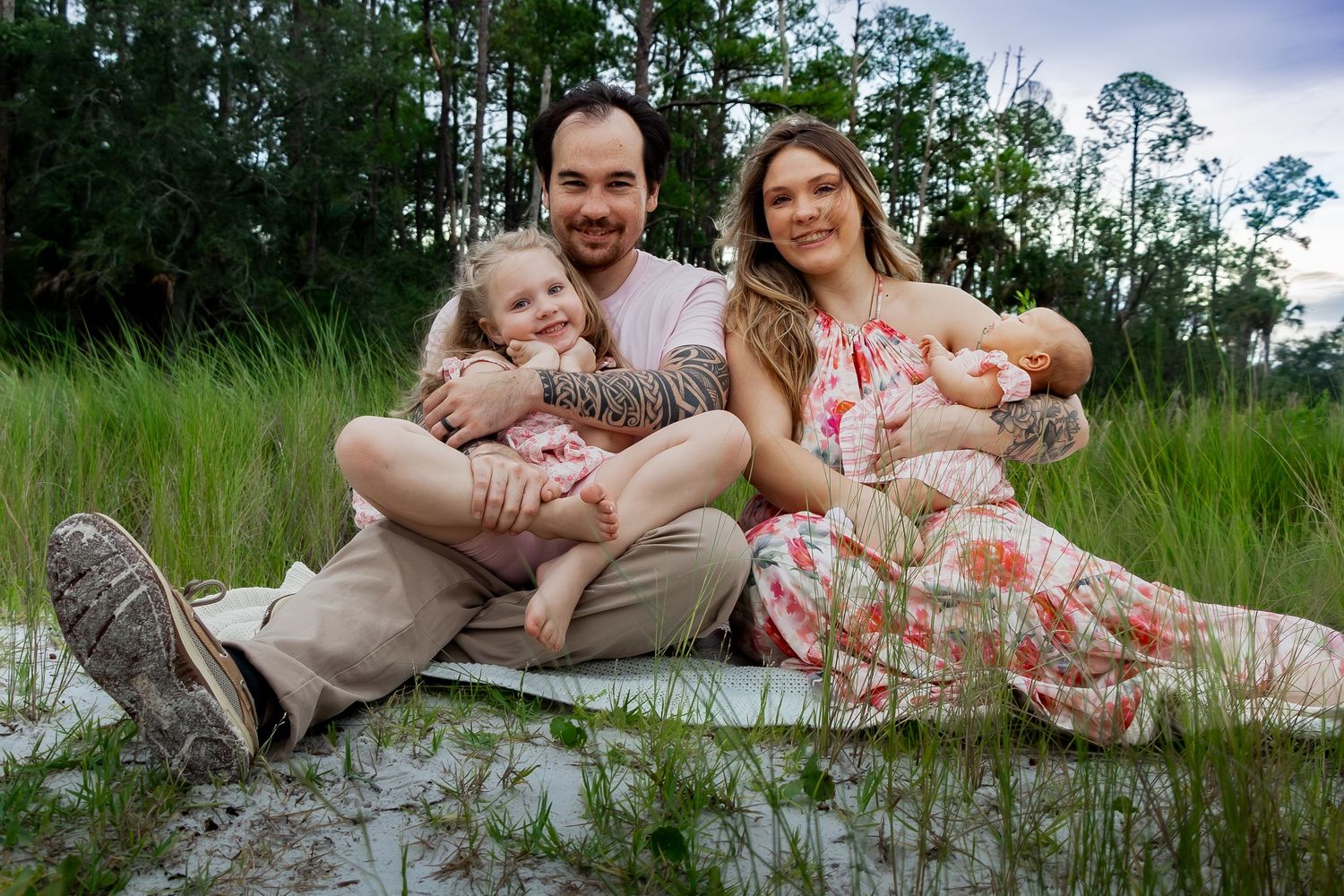 Happy family of four sitting in green meadow at dusk, parents holding young children outdoors.
