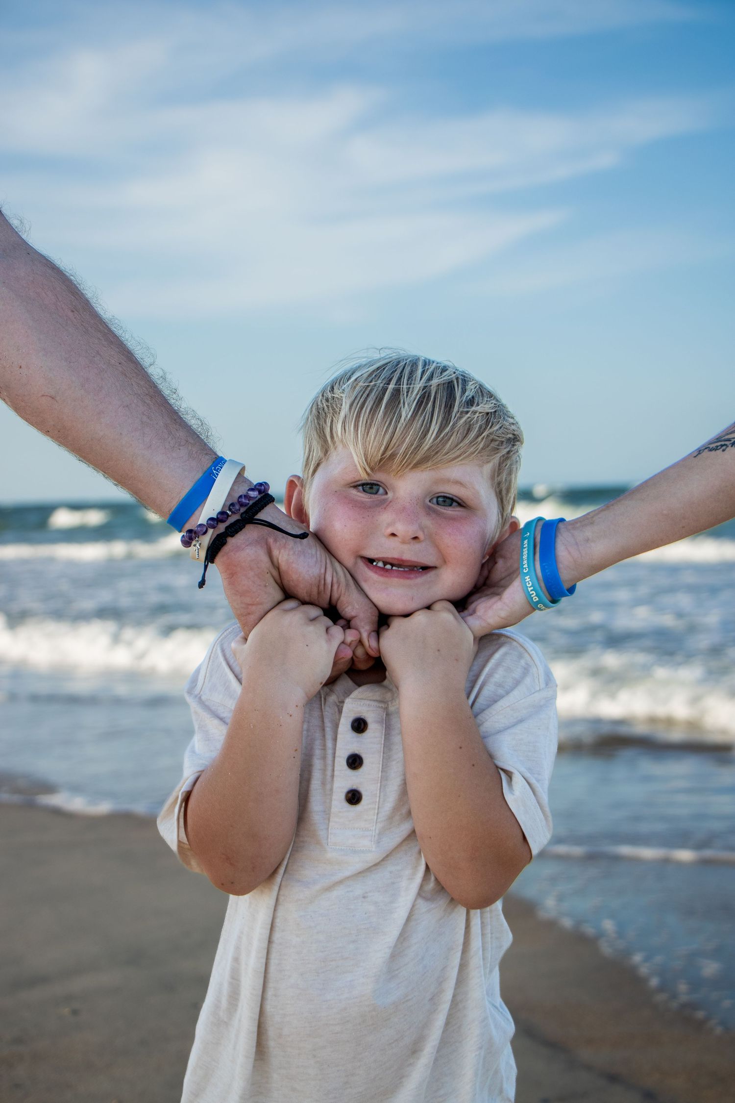 A childhood portrait of a boy holding his parents hands on the beach with the waves and sand in the background.