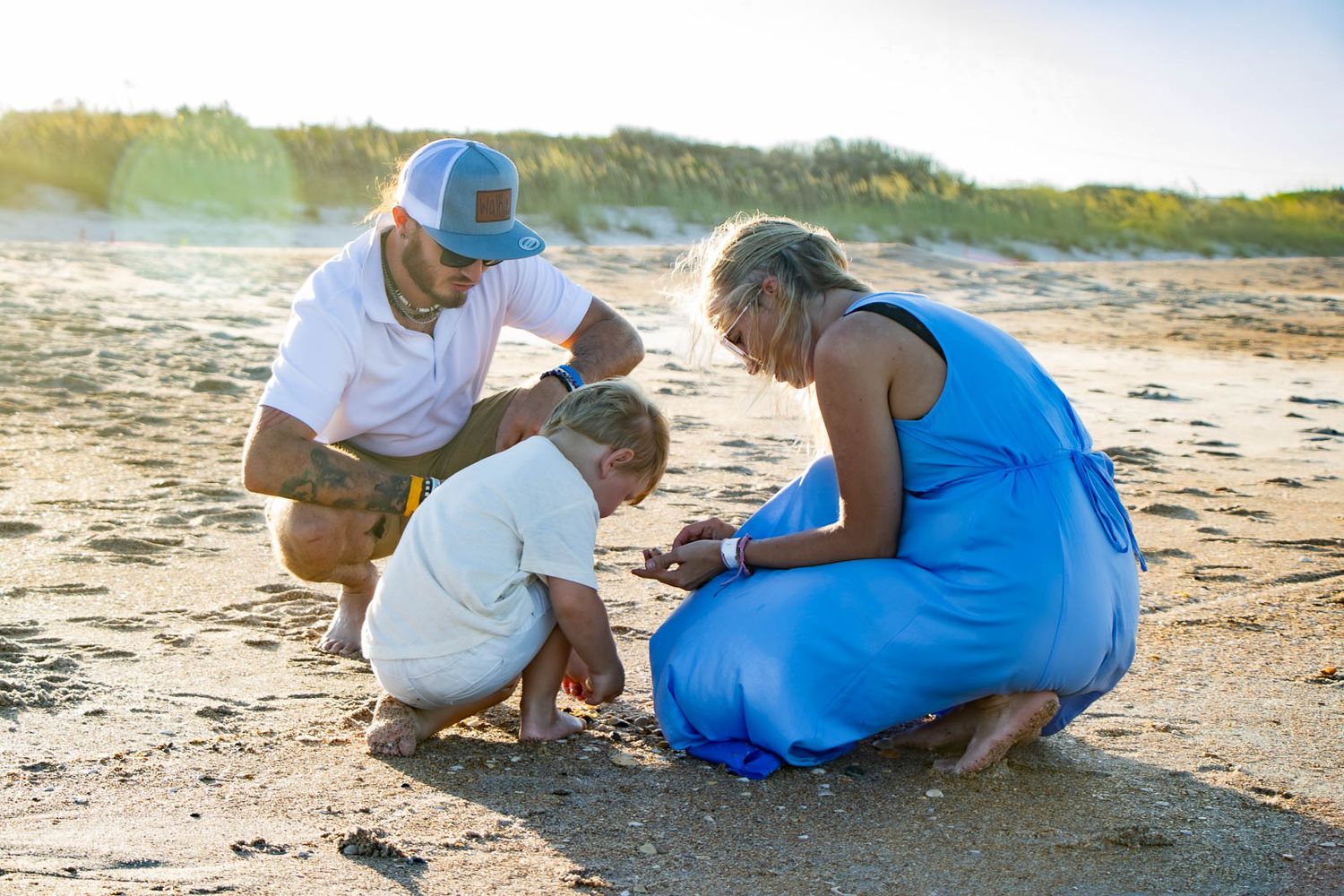 Family of three examining something in the sand on a sunny beach, with golden light and dunes in the background.