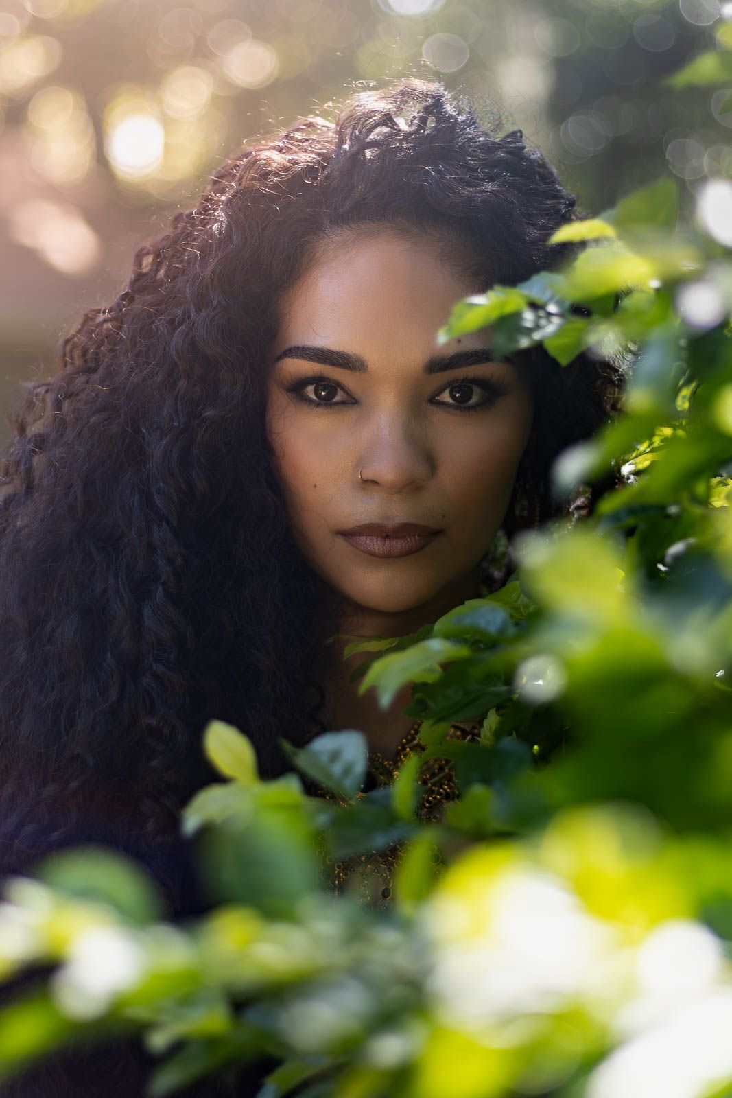 A young woman with long curly hair peers through lush green leaves in a sunlit outdoor setting for a portrait session in Jacksonville, fl.