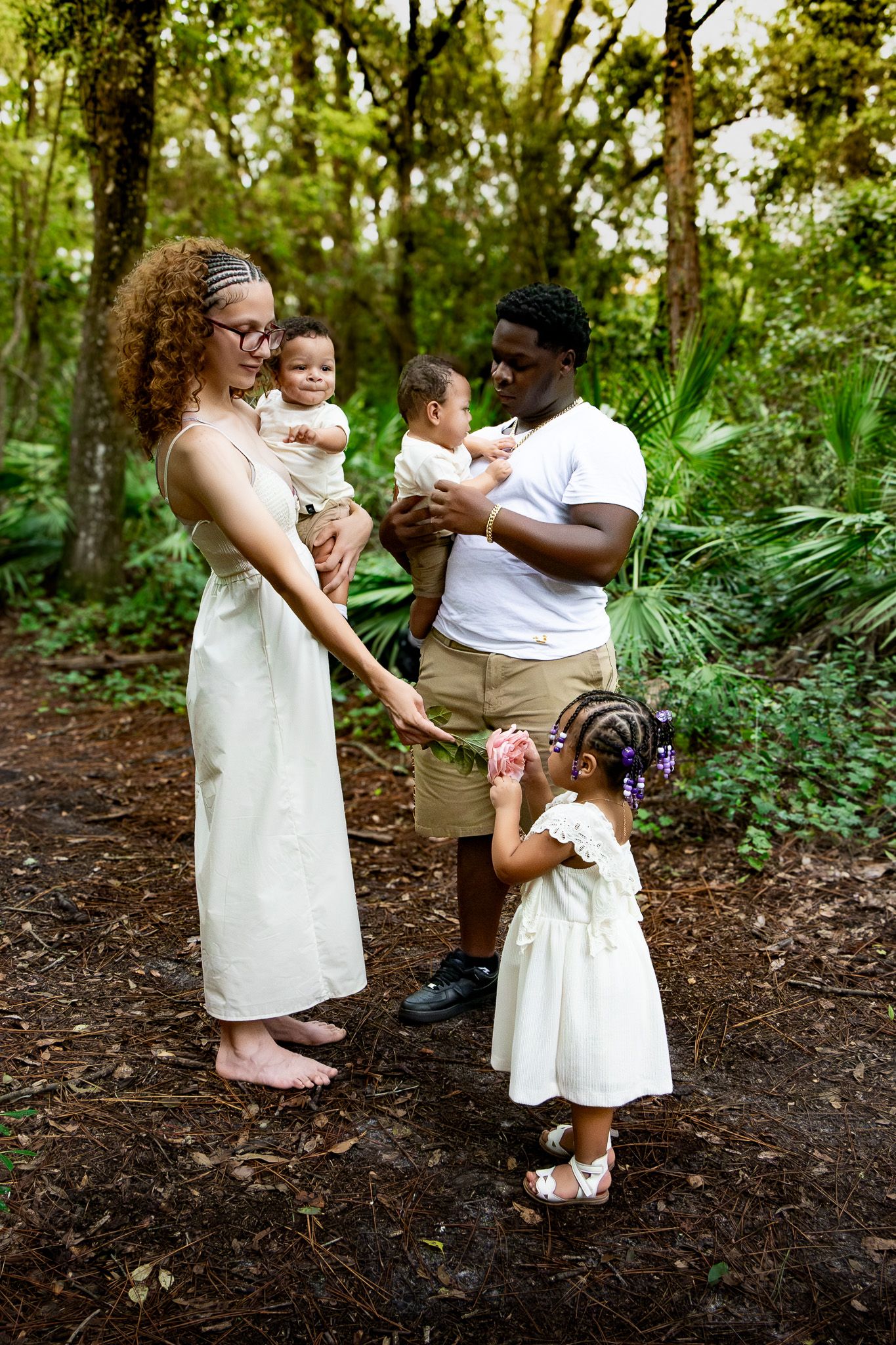 A family of four poses in a lush forest, parents holding babies while a toddler girl stands between them.