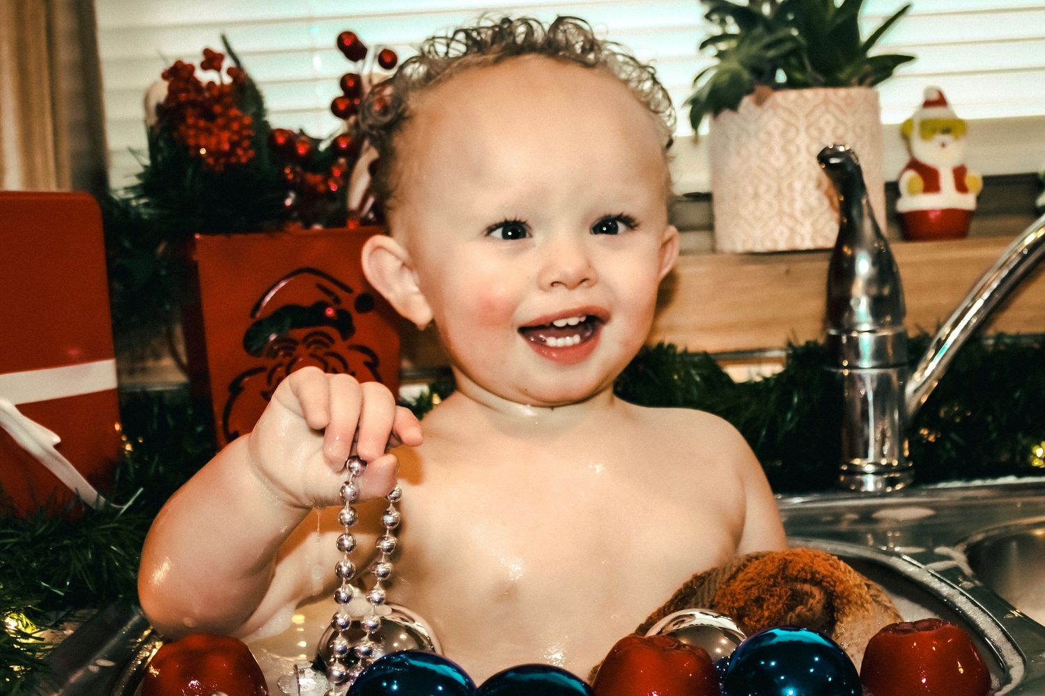 A baby during a childhood milestone shoot sitting in the sink playing with Christmas tree ornaments.