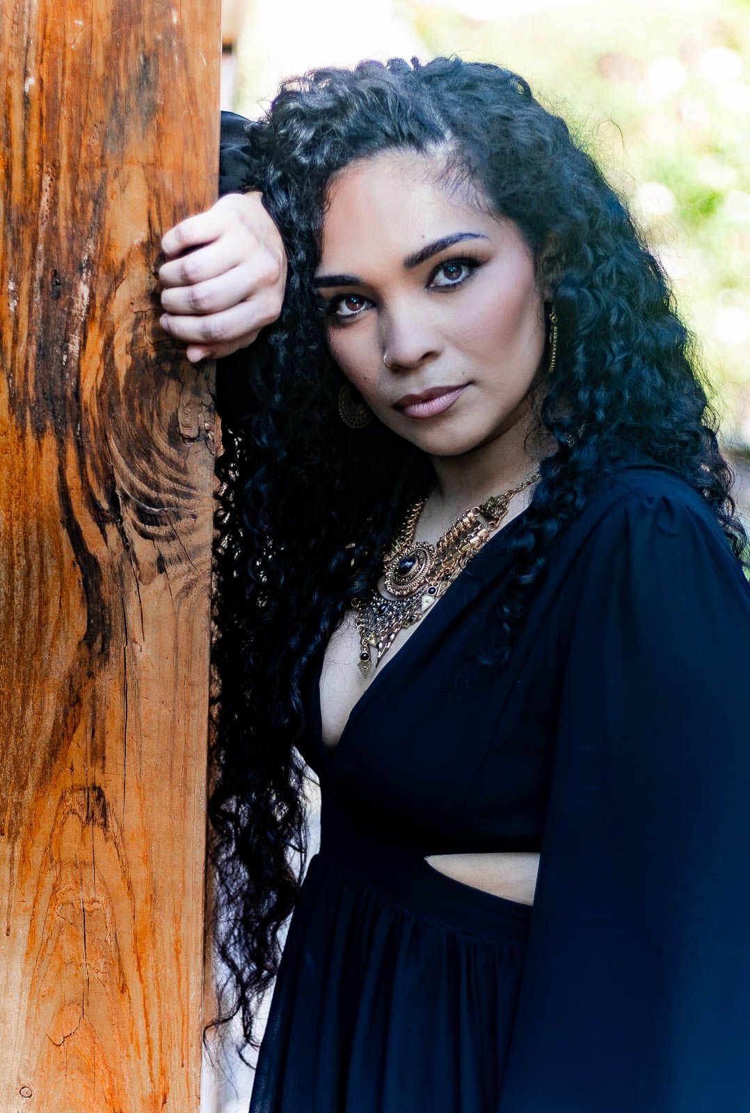Woman with curly black hair in dress leaning against wooden post, wearing gold necklace during a photography portrait session in floria.