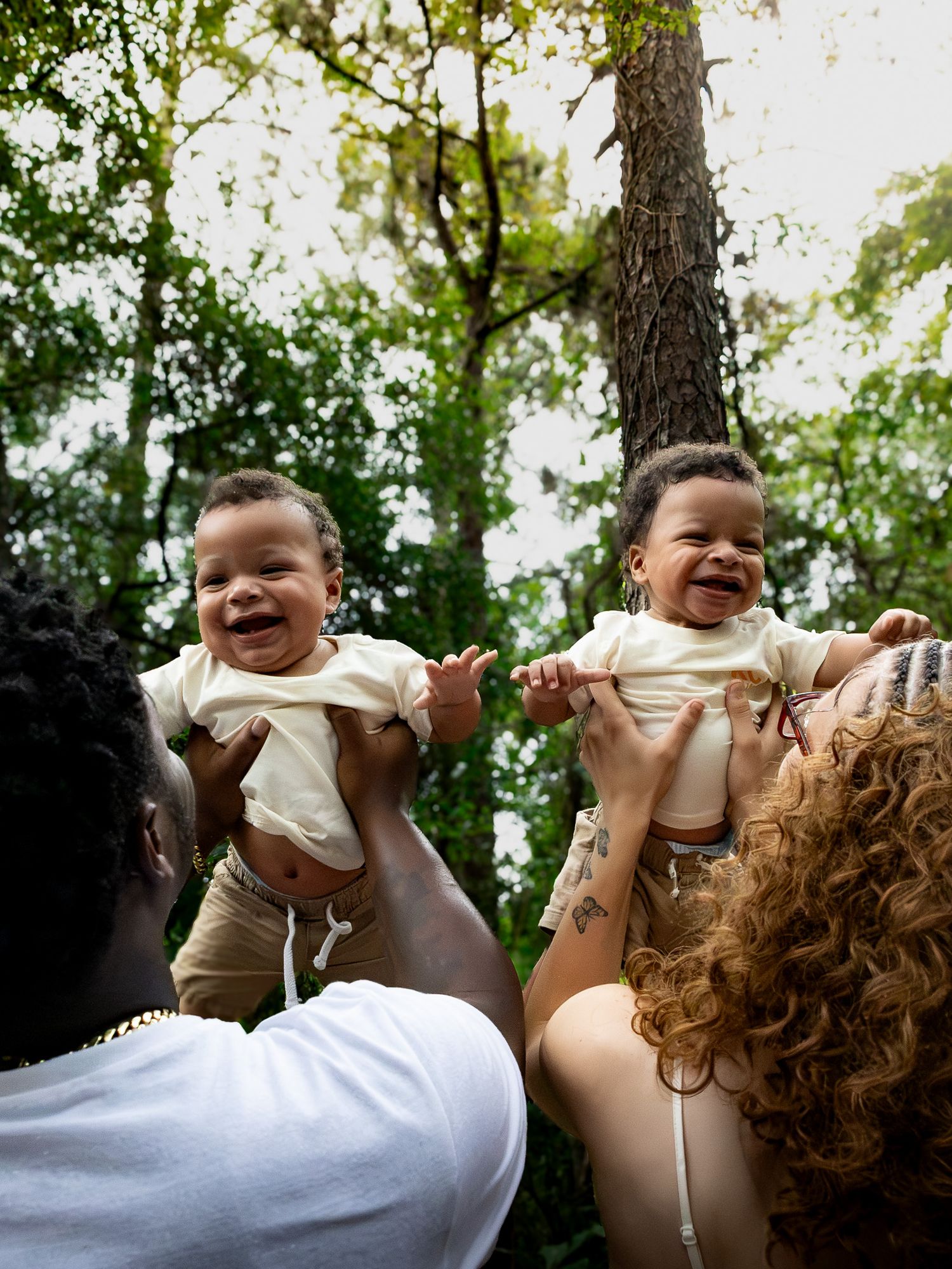 Two parents joyfully lift smiling twin babies outdoors among lush green trees in a heartwarming family moment.