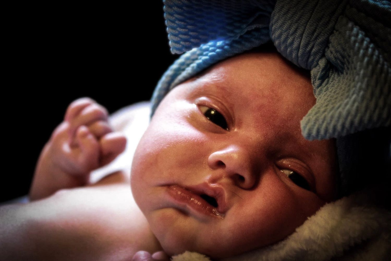 A newborn baby girl wearing a big blue bow looking at the camera with her fist balled up and a black background.