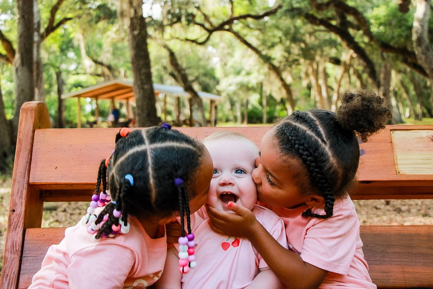 Three children sitting on a bench with a baby in the middle. Bright pink and green colors with trees in the background.