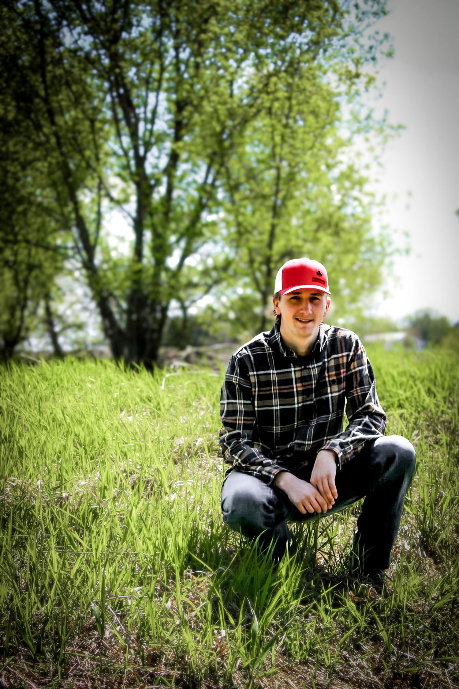 A boy kneeling in the grass posing for his senior portraits in the woods.