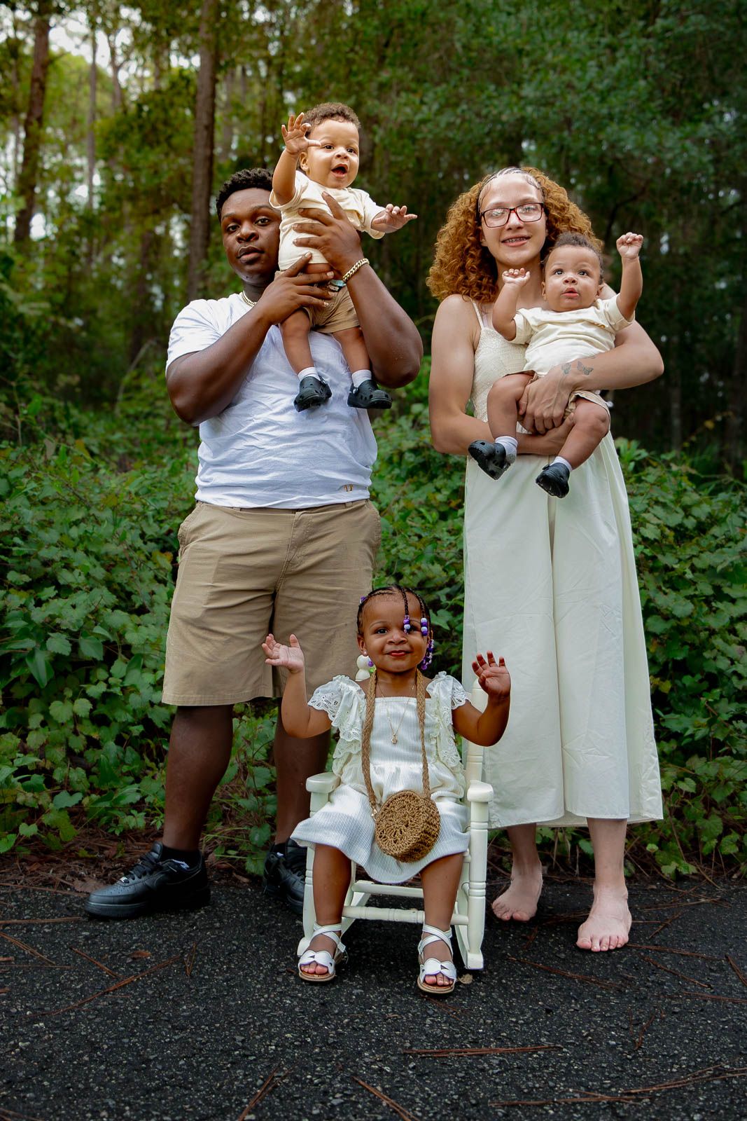 A joyful family posing outdoors, with two adults holding babies and a toddler sitting in a small white rocking chair for a family portrait.