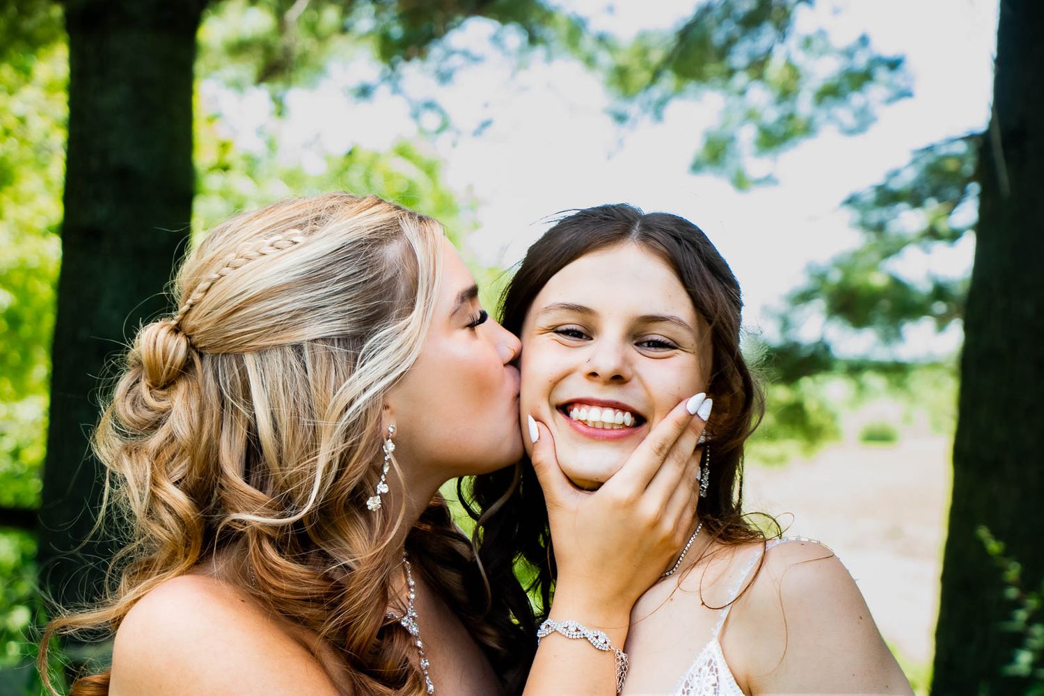 Two young women in formal attire share a sweet kiss on the cheek outdoors, smiling joyfully.