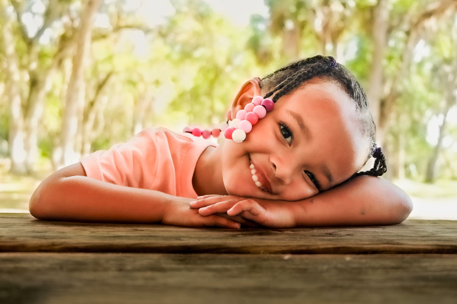 Smiling young girl resting her chin on a wooden picnic table outdoors, wearing a pink top and floral hair accessories.