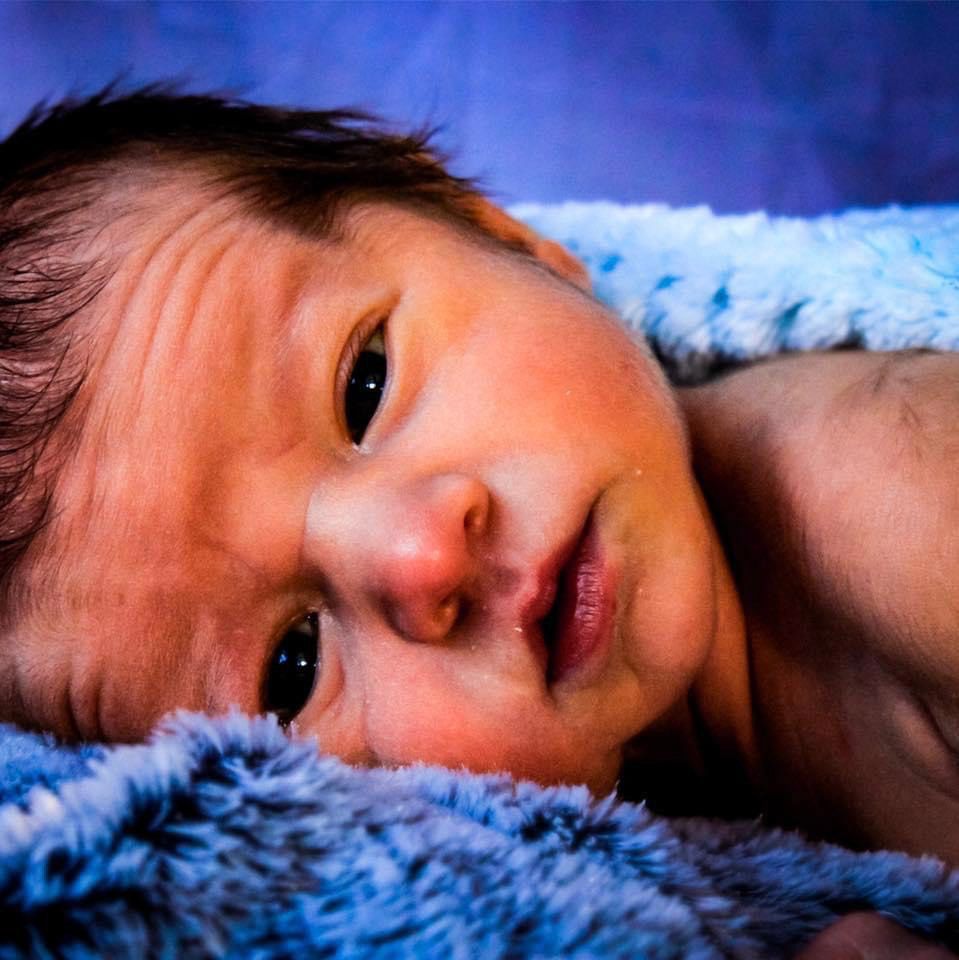 A newborn looking and smiling into the camera with a blue background.