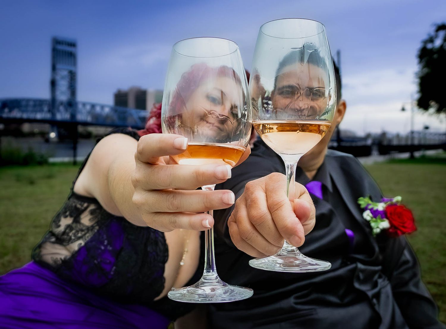 Couple toasting with wine glasses outdoors, faces reflected in glasses, bridge in background at dusk.