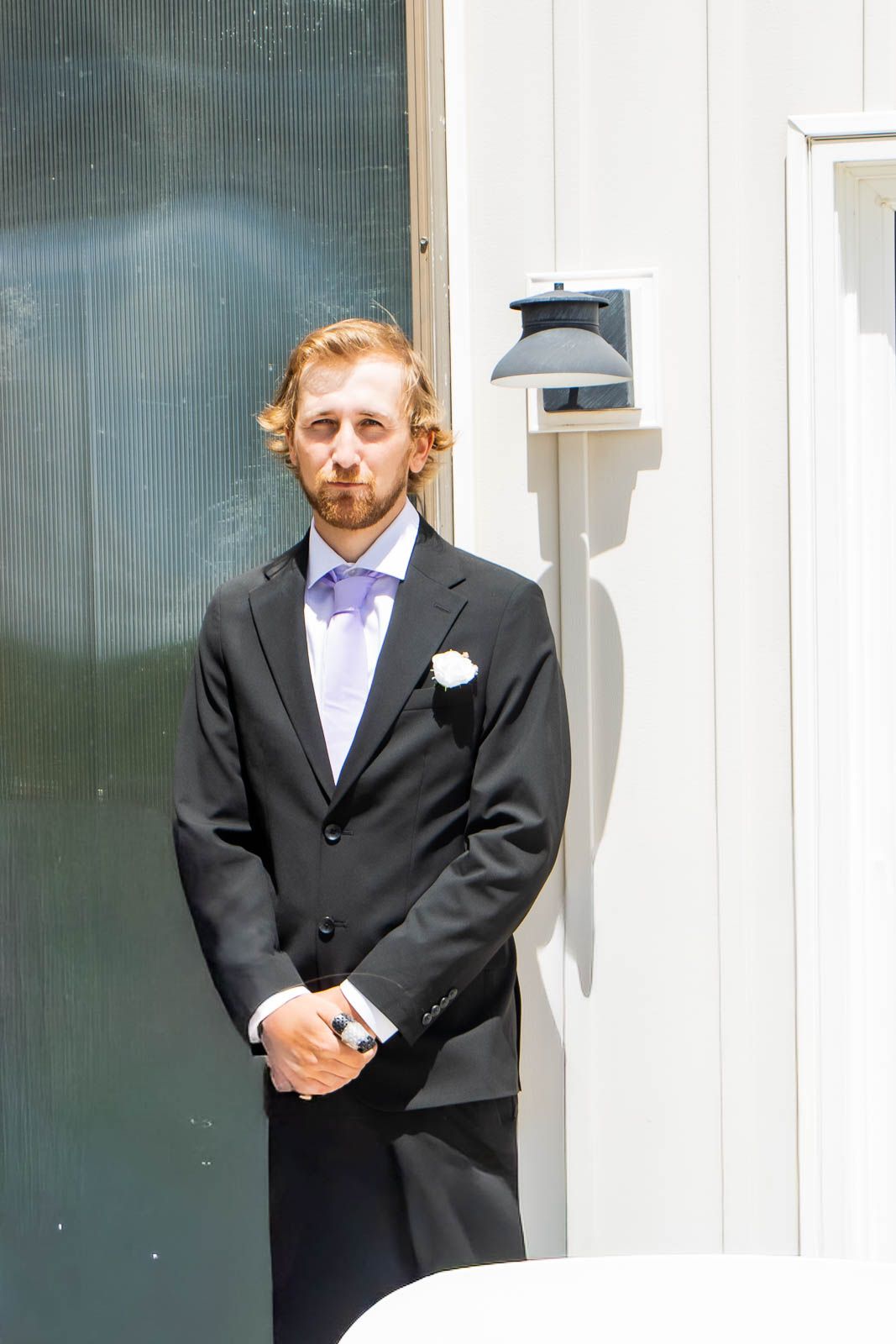Groom in black suit with white boutonniere and lavender tie stands outdoors on wedding day.